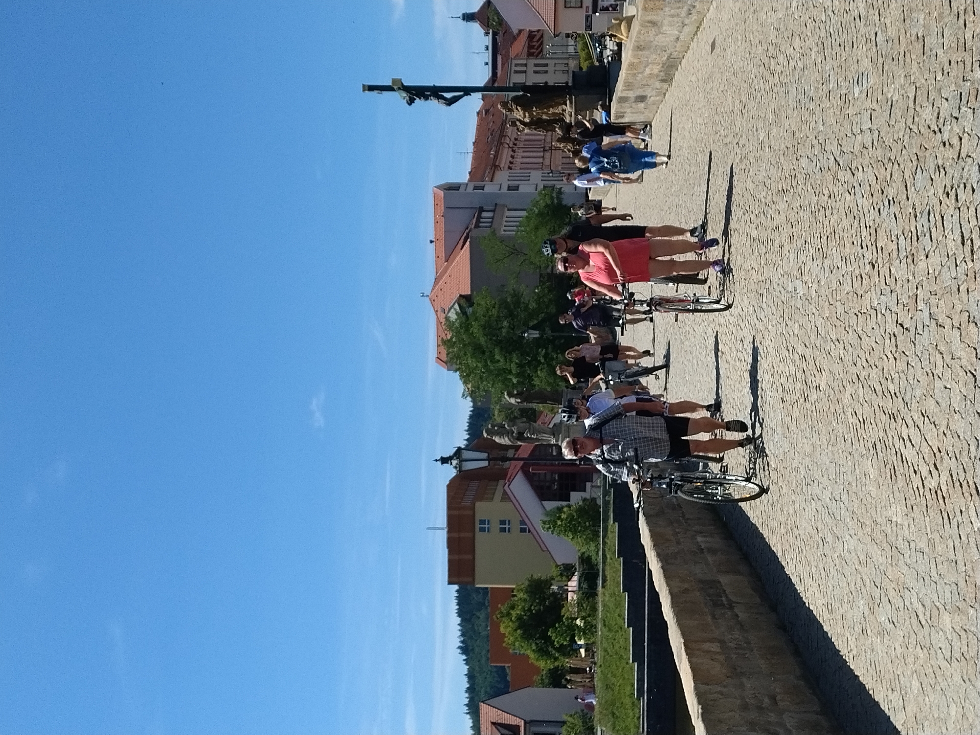 On a sunny day, a group of people are walking and cycling across a cobblestone bridge in Písek, Czechia. The bridge is lined with a stone wall. In the background, buildings with red-tiled roofs and trees are visible. A statue of Jesus on the cross stands on the right side of the bridge.