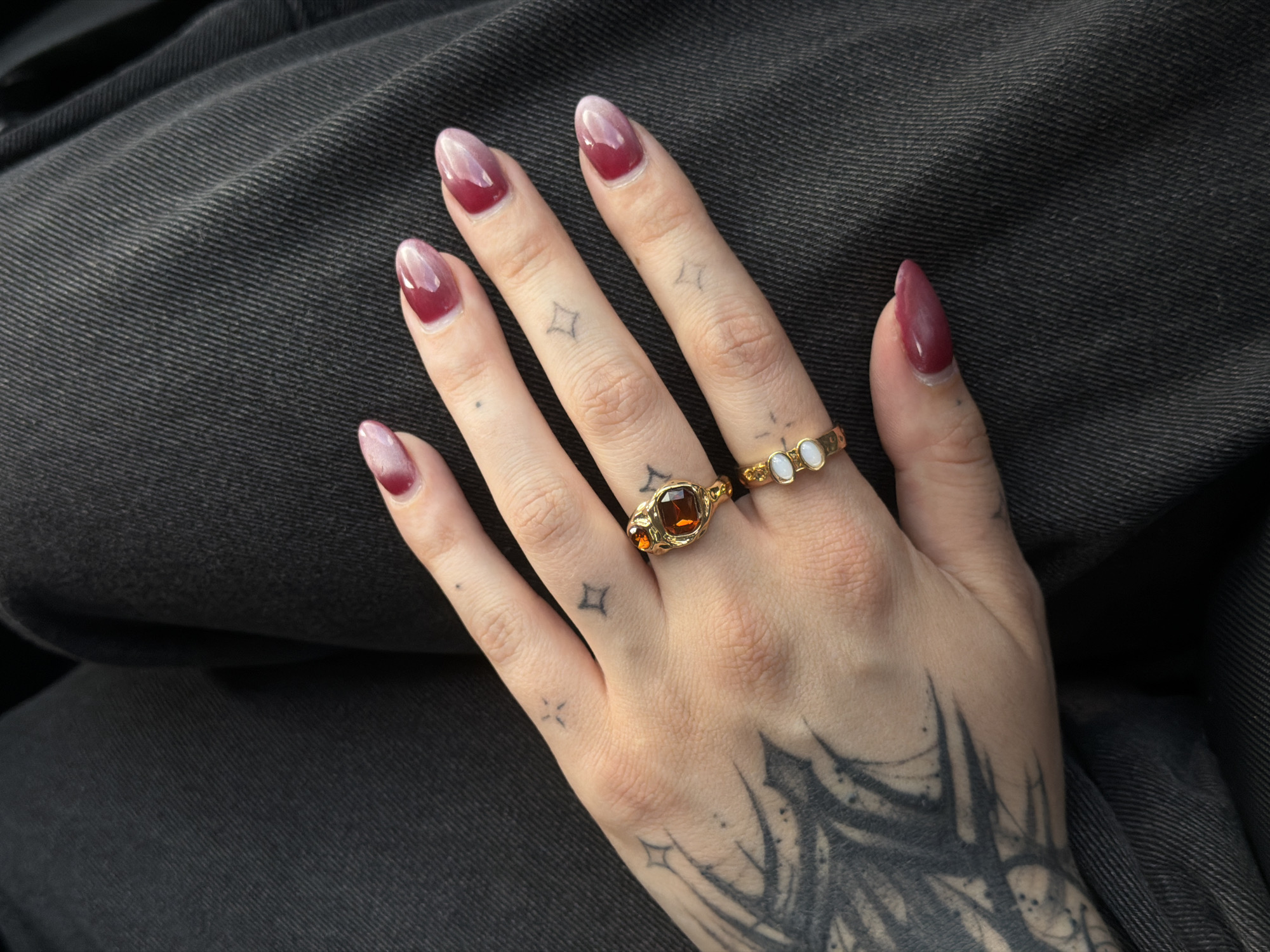 A close-up, overhead shot shows a left hand resting on dark gray denim fabric. The hand has several small tattoos, including stars and a larger, dark design on the side of the palm and wrist area. The fingernails are painted with a gradient of deep red at the tips fading to a lighter pink towards the base. Two gold rings are worn on the fingers. One ring on the middle finger has a large, faceted amber-colored stone. The other ring, on the ring finger, has small white oval stones. The denim fabric appears to be jeans or pants. The lighting suggests an outdoor or well-lit indoor environment. There is no indication of the location being in a city.