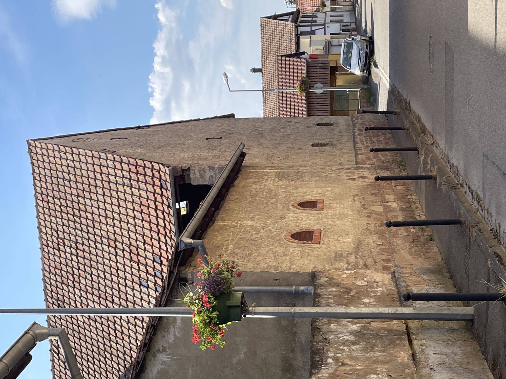 The image shows a street scene in Gerstheim, France, likely taken during the daytime on a clear, sunny day with some clouds. The focus is on the side of an old stone building with a red-tiled roof. The building has two small, arched windows near the top of its wall and a hanging planter filled with colorful flowers, including red and purple blooms, attached to a pipe.
In the background, there are more buildings with red-tiled roofs, some of which appear to be houses or shops. A street runs along the right side of the image, bordered by a low wall and several black metal posts. A silver car is parked on the left side of the street. A street lamp is visible on the left side of the image, extending over the street. The overall impression is a quiet, residential or semi-commercial street in a European town.