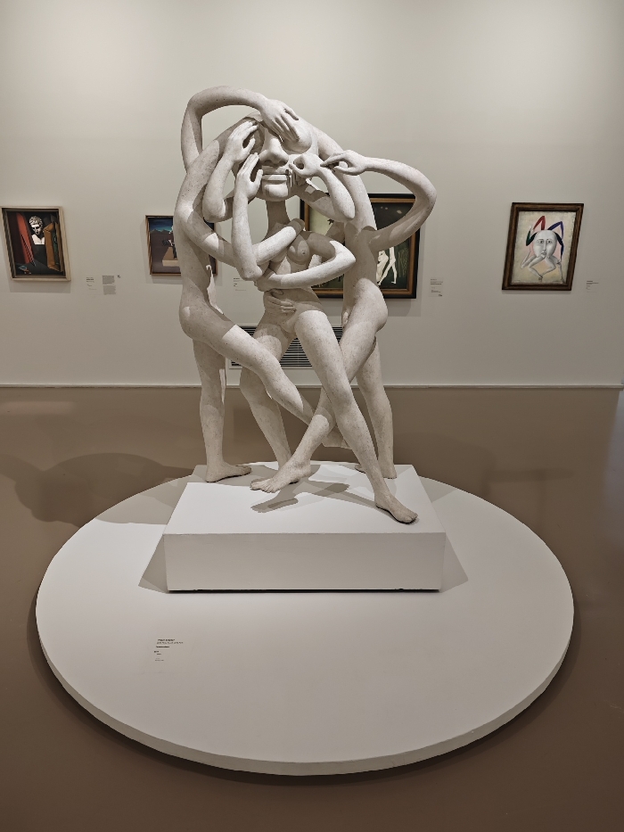 The image captures a detailed view of a large, complex sculpture displayed within a museum gallery, specifically the Centre Pompidou in Paris, France.
The central subject is a prominent, light-colored gypsum sculpture titled "The Scream" (Le Cri) by Germaine Richier, created in 1957. It depicts multiple intertwined, emaciated, nude humanoid figures in a dynamic and distressed arrangement. A central figure's face is obscured by hands, while other hands pull at its jawline, creating an expression of intense anguish. Surrounding figures, with elongated limbs, are interwoven with the central form, suggesting embrace, struggle, or emergence. The sculpture possesses a raw, textured surface and rests on a white rectangular plinth, which is centered on a larger white circular platform.
The gallery environment features off-white walls and a smooth, light brown floor. Several framed paintings are visible on the walls in the background, suggesting a collection of modern art, possibly surrealist or expressionist in style. These include a portrait on the far left, a darker composition mid-left, and a distinct portrait-like piece with a stylized figure and headwear on the far right. Overhead lighting illuminates the scene, casting soft shadows beneath the sculpture and display platform. Small descriptive labels are visible on the walls adjacent to the paintings and on the circular platform, providing contextual information about the artworks.