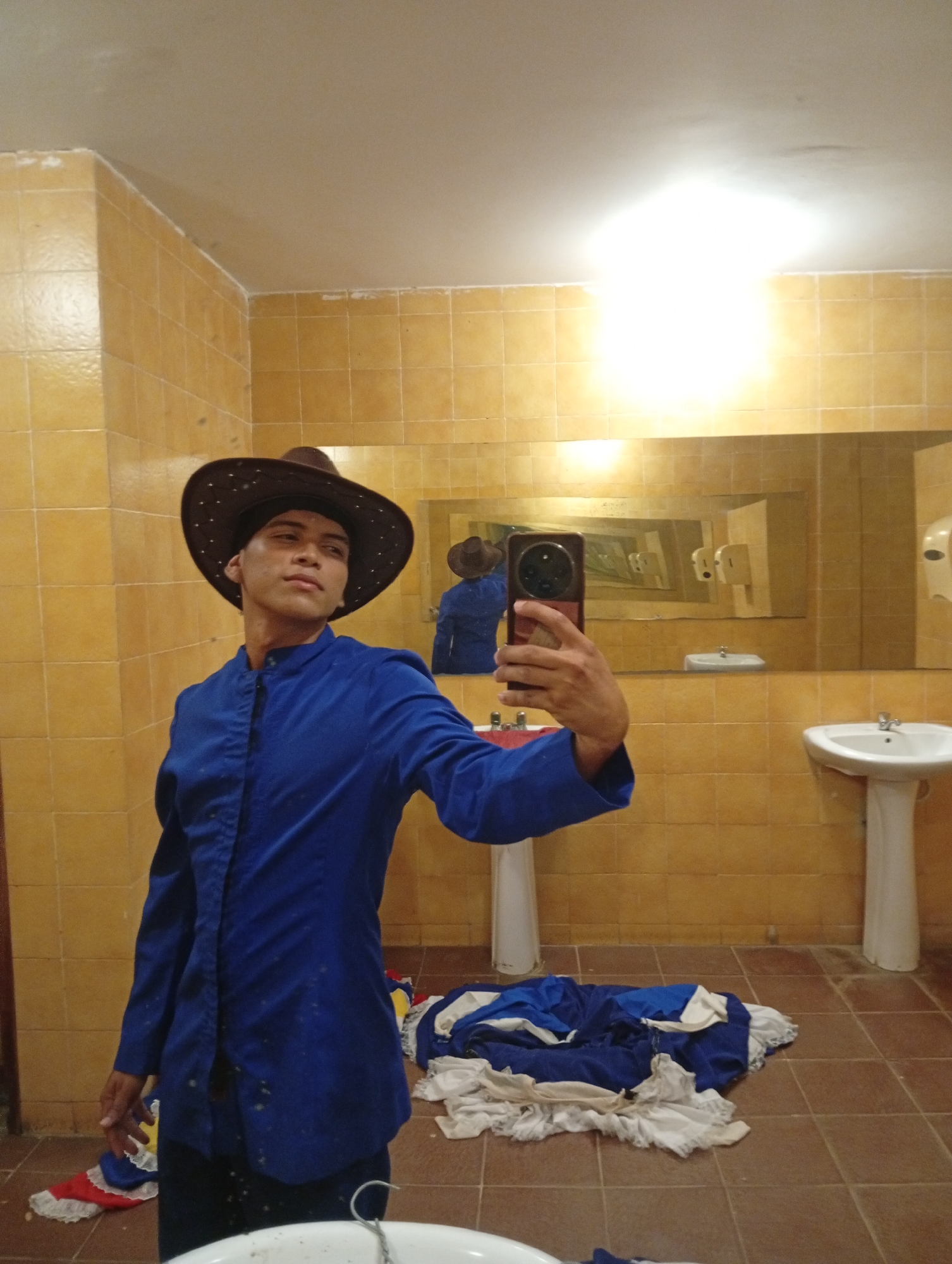 A young man is taking a selfie in a bathroom. He is wearing a brown cowboy hat and a long-sleeved blue shirt. He is holding a cell phone in his right hand. The bathroom has yellow tiled walls and a brown tiled floor. There are two sinks, one on either side of the man. There is a mirror behind him, and a pile of clothing on the floor. The location is Pedro González, Venezuela.