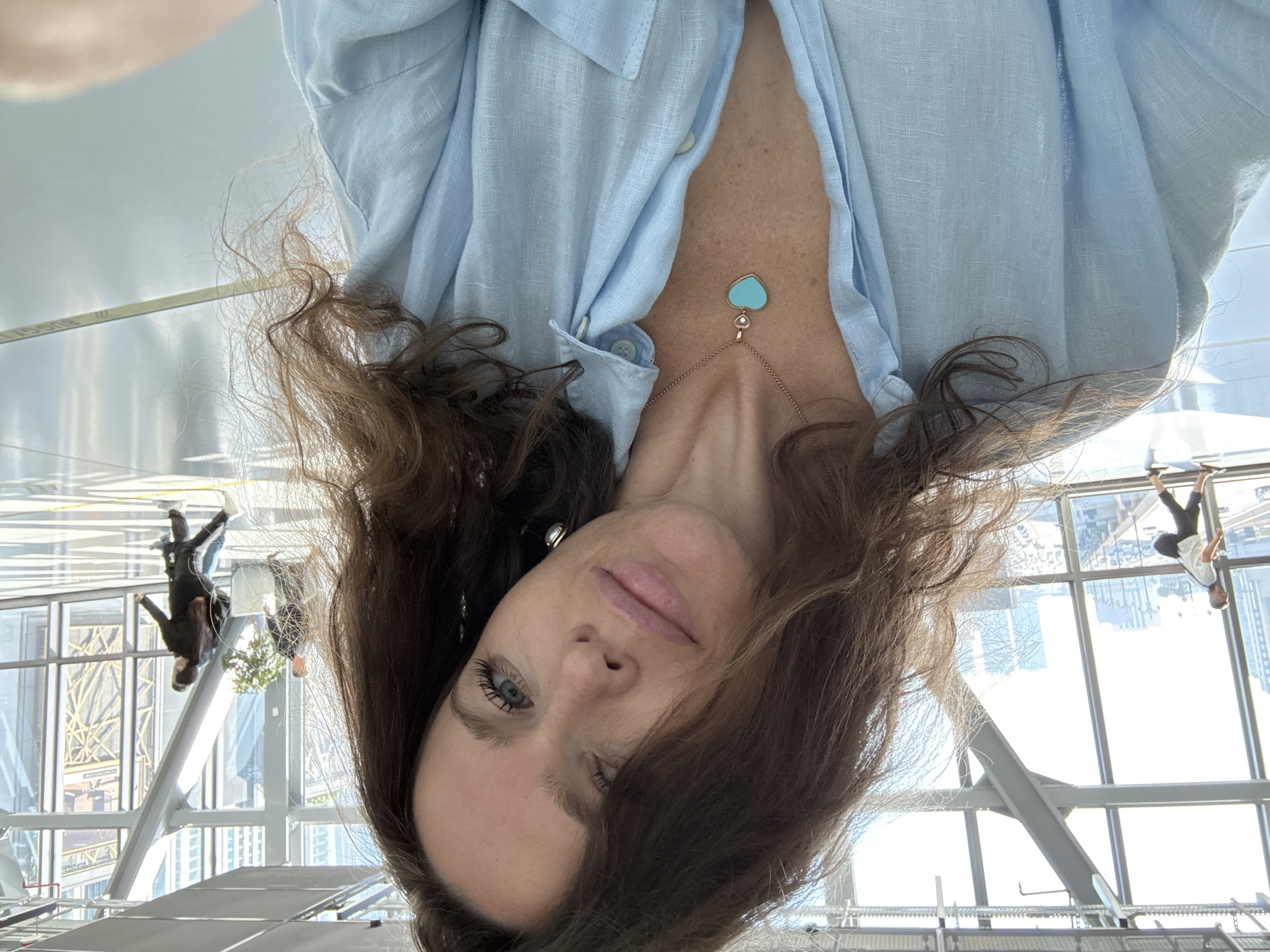 A woman takes a selfie in a modern, sunlit indoor walkway with large glass windows offering a view of a cityscape. She has long, wavy brown hair and is wearing a light blue collared shirt and a necklace with a heart-shaped pendant. In the background, people are walking in a brightly lit space with polished floors. The architecture suggests an airport or a large public building. This location is in Dubai, United Arab Emirates.