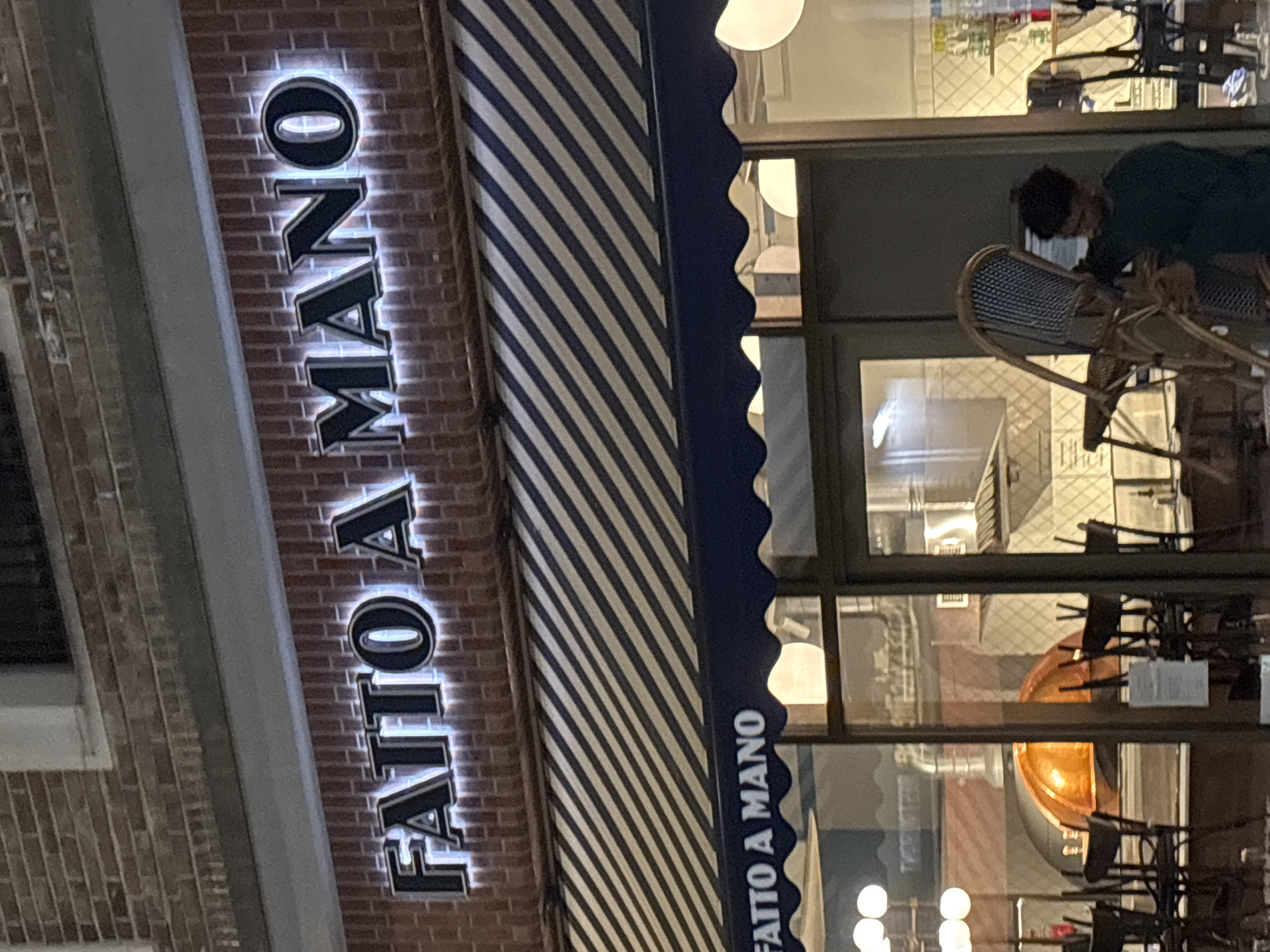 The image shows the exterior of "FATTO A MANO," a restaurant in London, United Kingdom. The sign, illuminated and vertical, is affixed to a brick facade next to a blue-and-white striped awning. The awning's edge is scalloped. Reflections in the glass windows reveal interior details, including chairs, tables, and a light fixture. A person is visible at a table outside, wearing a mask.