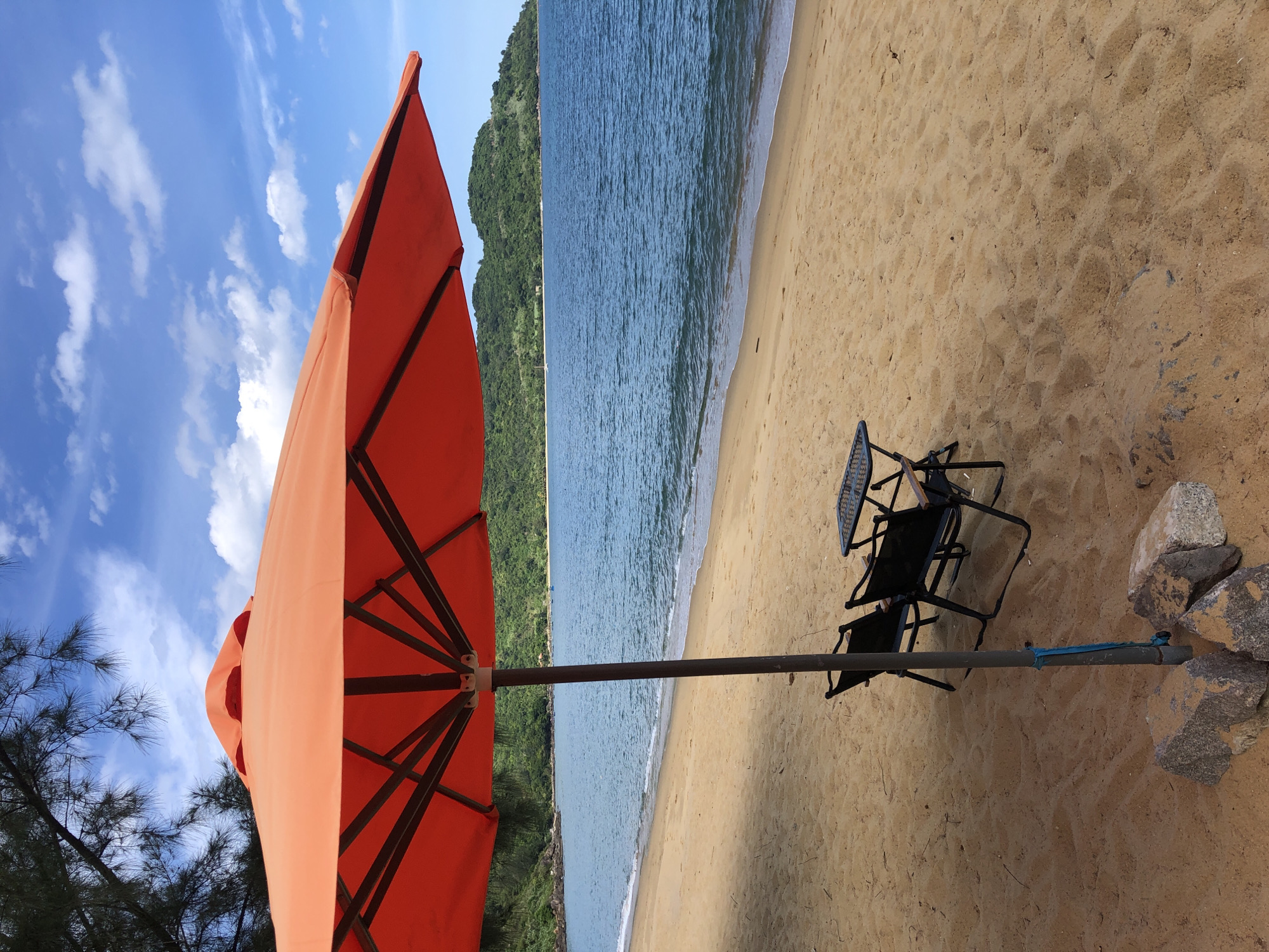 This is a photograph of a deserted beach with an orange umbrella casting a shadow over two empty chairs and a small table. The sand is a warm, golden color, and the ocean is a calm, deep blue. In the distance, a lush green hillside slopes down to the water. The sky above is a bright blue, dotted with fluffy white clouds. The scene evokes a sense of peace and relaxation. The location is likely a beach in Da Nang, Vietnam, known for its beautiful coastlines and tropical scenery.