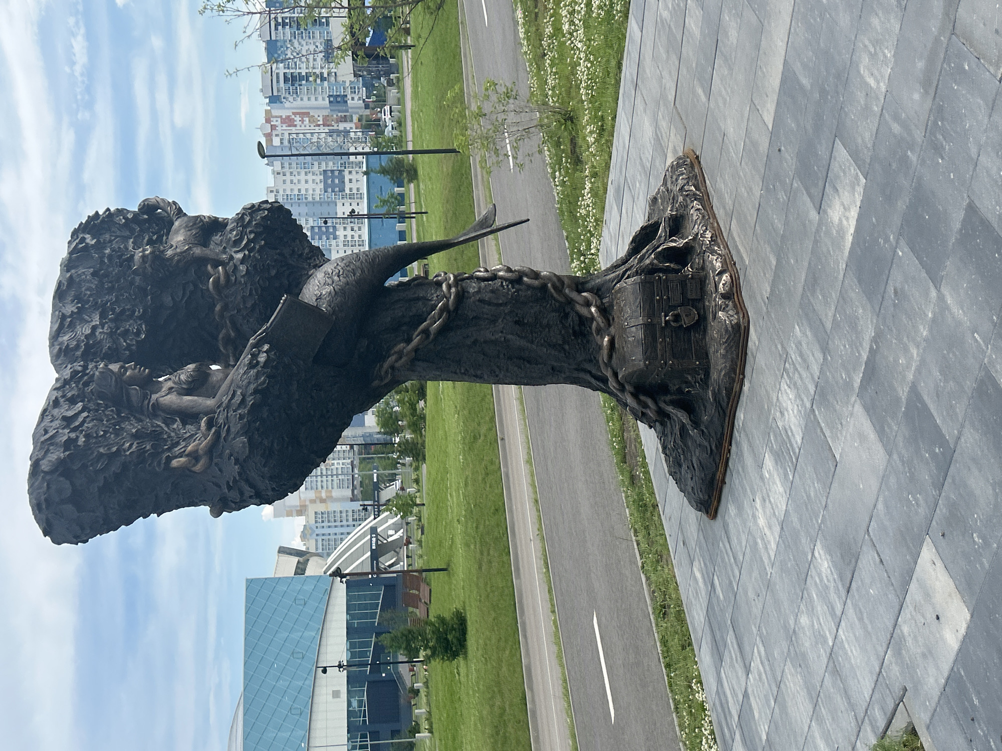 A bronze sculpture depicting a mermaid entwined with a tree stands on a paved area in Kemerovo, Russia. The sculpture features a mermaid figure with a tail and human upper body, seated amongst stylized tree branches. A small chest with a lock is positioned at the base of the sculpture, and chains wrap around the tree trunk. In the background, a modern building with a blue glass facade and a cityscape of residential buildings are visible. The scene is set on a bright, cloudy day, with a road and a grassy area with scattered white flowers in the foreground.