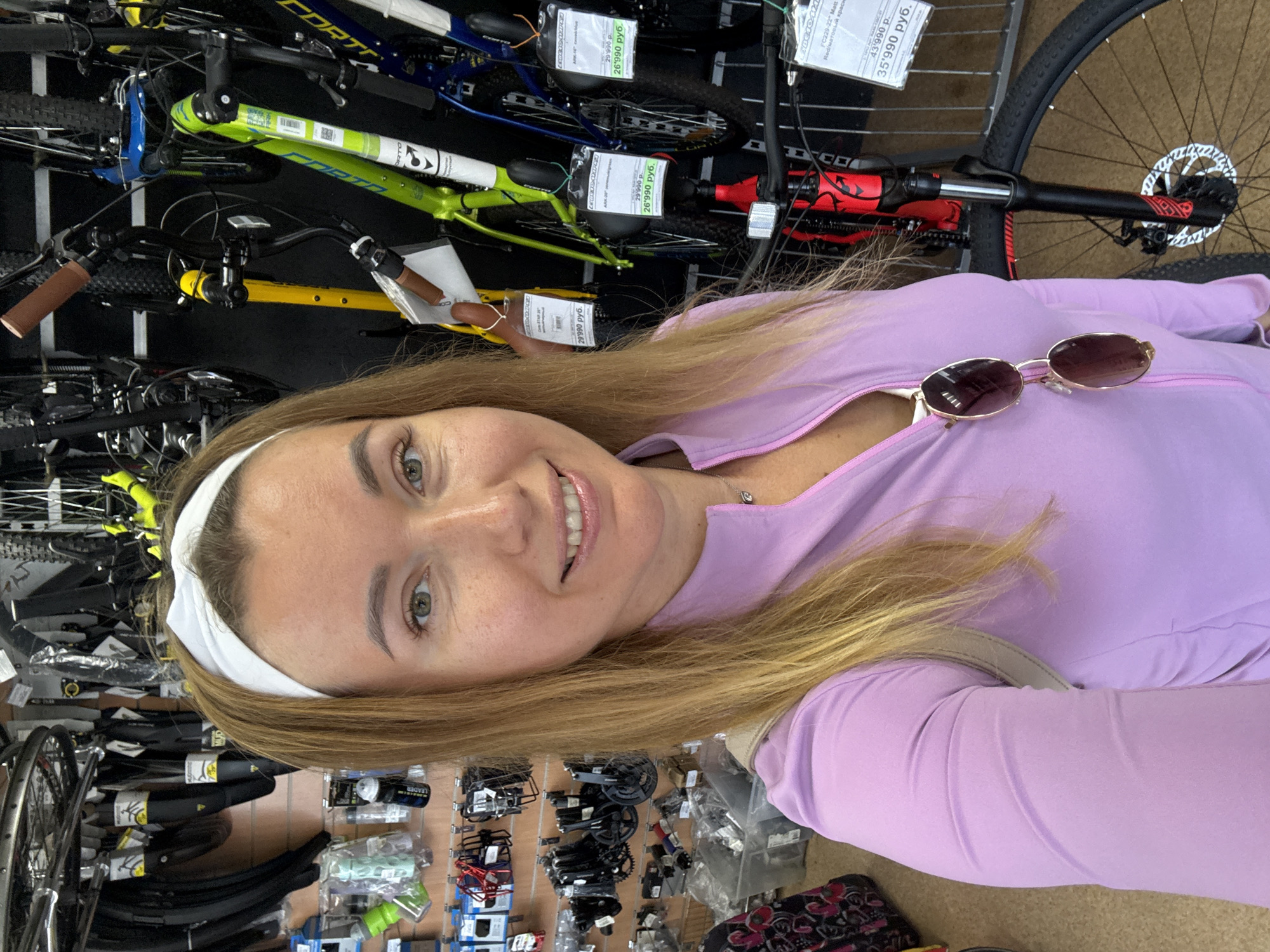 A woman with blonde hair and a white headband takes a selfie in a bicycle shop. She is wearing a pink zip-up jacket and sunglasses are hanging from the zipper. In the background, various bicycles and bicycle parts are displayed on shelves and racks. The shop appears to be well-lit and organized. The woman's face is partially visible as she smiles at the camera.