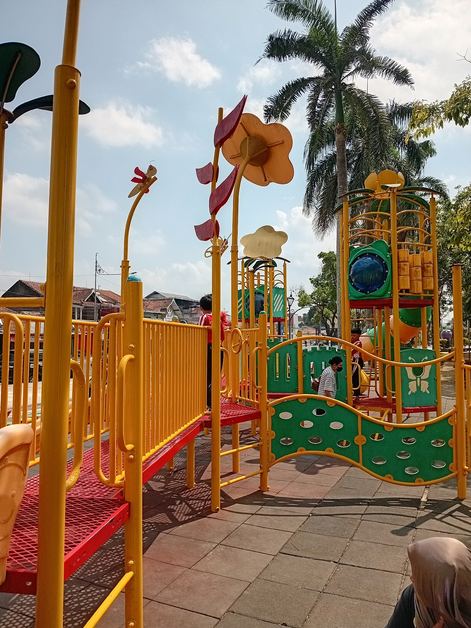The image shows a colorful playground with yellow and green structures. There are several children present, some of whom are playing on the equipment. The playground is situated in an outdoor area with trees and buildings visible in the background. The ground is covered with interlocking rubber tiles. The sky is partly cloudy. The architecture of the buildings in the background, particularly the tiled roofs, suggests a location in Indonesia, consistent with Purworejo. One person is wearing a headscarf, which is common in Indonesia. The presence of palm trees further supports this regional identification.