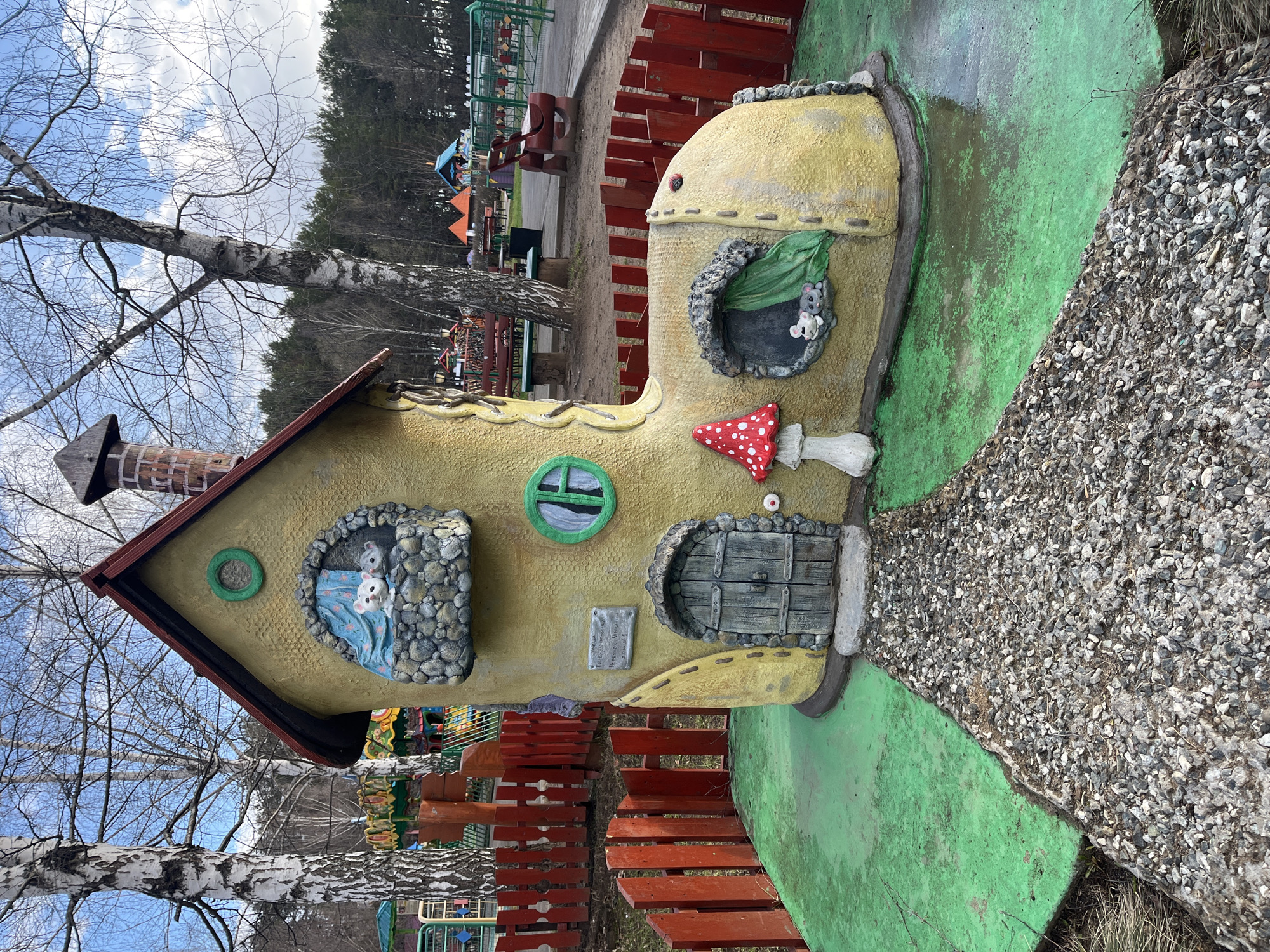 The image displays a whimsical children's play area, dominated by a large, yellow shoe-shaped playhouse designed like a house. The structure features a brown and reddish pitched roof with a brick chimney. It has multiple windows: a circular green one, another with a stone-like frame revealing two white mice sleeping soundly under a blue blanket, and a third with a leaf-like frame where two grey mice peek out. A rustic, wooden-style door, also framed with stone, serves as the entrance, with a red and white spotted mushroom decorating the exterior. A path made of small grey stones leads up to the door, bordered by green painted ground. A small plaque next to the door bears Cyrillic text, which reads "HERE LIVES GRANDFATHER FROST."
The setting is an outdoor park or playground. In the background, various colorful playground equipment, including red wooden fences, slides, and other structures, are visible. The natural surroundings include tall evergreen trees in the distance and bare birch trees in the foreground, indicating either late winter or early spring. The sky is partially cloudy with patches of blue, suggesting a bright daytime. No people are present in the scene. The overall atmosphere is one of playful fantasy and imagination, typical of a children's attraction in Chaikovsky, Russia.