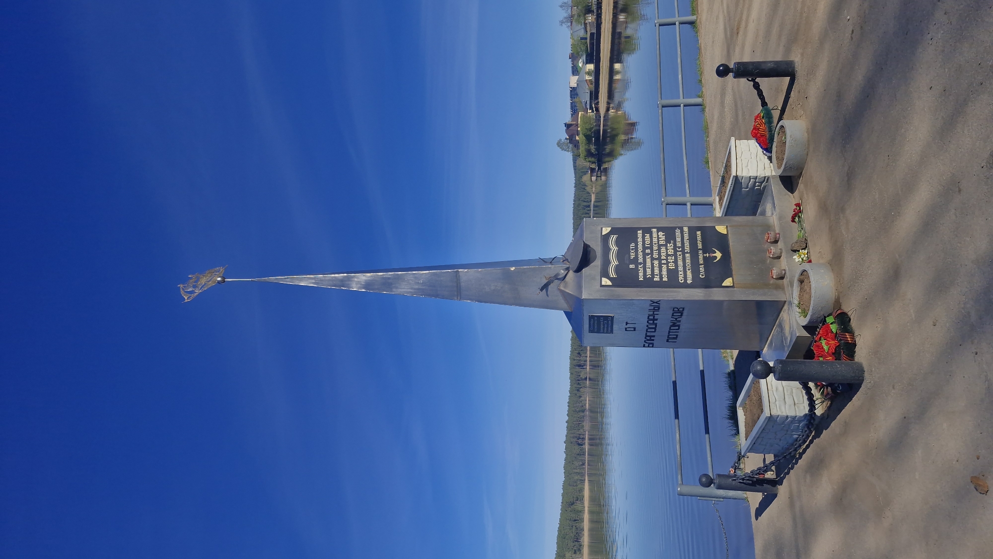The image captures a monument in Otchor, Russia. It features a silver, triangular spire atop a cubic base. The base has inscriptions in Russian, including "FROM GRATEFUL DESCENDANTS" and a dedication to young volunteers who fought during the Great Patriotic War (World War II). The monument is decorated with flowers and wreaths. It's situated on a paved area near a body of water, possibly a river or lake, with a reflection of trees on the horizon. The sky is a gradient of light to dark blue. A small flag or symbol is present at the very tip of the spire.