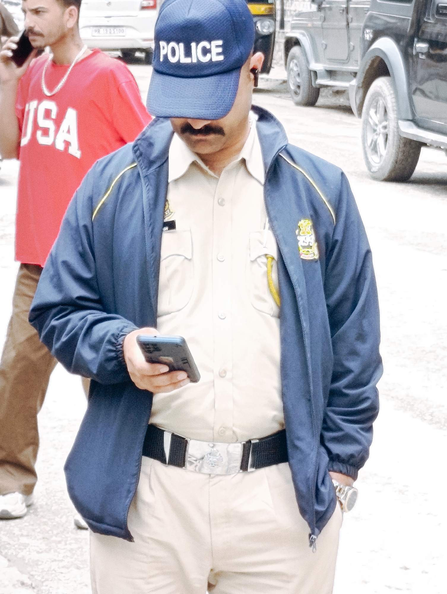 A man wearing a blue police cap with "POLICE" printed in white, a beige uniform shirt, and a dark blue jacket with yellow trim is holding a dark smartphone in his left hand. He has a mustache and is looking down. A black belt with a silver buckle is visible around his waist. He is wearing light-colored pants. To his left, another man in a red t-shirt with "USA" printed in white is visible, talking on a mobile phone. In the background, several vehicles, including what appears to be a black SUV and a three-wheeled auto-rickshaw, are parked on a paved surface. The overall scene suggests an outdoor urban or semi-urban environment.