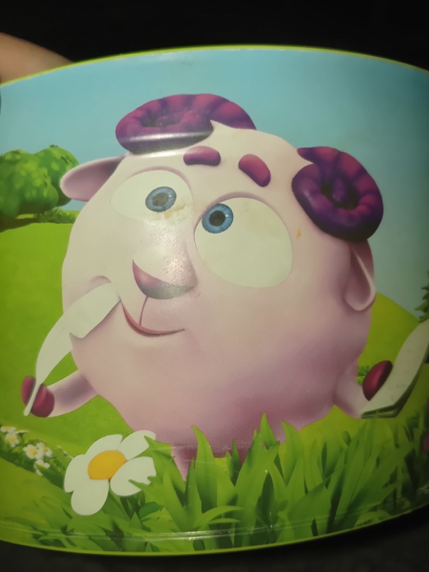 The image depicts a cartoon character resembling a sheep with pink skin, blue eyes, and purple curled horns. It has a white patch over one eye and a white tongue sticking out. The sheep character is set against a backdrop of a green field with grass and a white flower with a yellow center. Behind the field is a green hill and a light blue sky.
There is no information in the image itself to indicate the city of Puchezh, Russia.