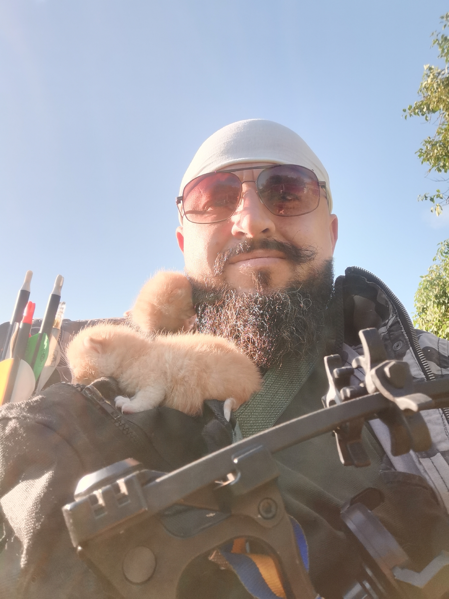 In Semypolky, Ukraine, a man with a beard and mustache, wearing sunglasses and a white bandana, is holding two small, light-colored kittens on his shoulder. He is also carrying a quiver of arrows with fletching visible, and a portion of a crossbow is in the foreground. The man is wearing a camouflage jacket over a green shirt. The scene appears to be outdoors, with a clear blue sky and some trees visible in the background.