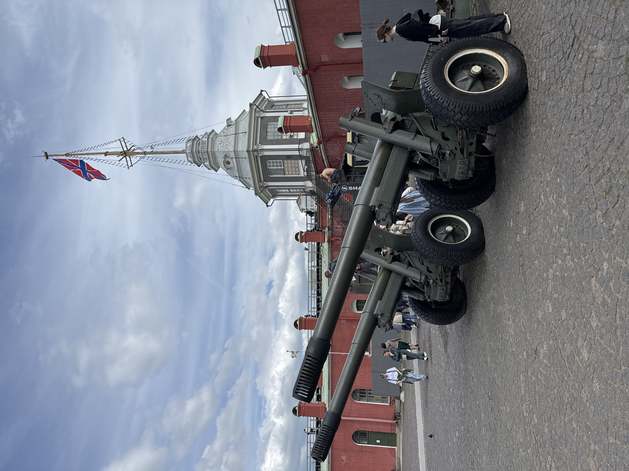The image depicts an outdoor scene at what appears to be the Peter and Paul Fortress in Saint Petersburg, Russia. In the foreground, several large, olive-green artillery pieces, likely historical cannons or howitzers, are prominently displayed on a cobblestone ground. These military ordnance feature robust black tires with white rims and long, dark barrels, some equipped with distinct muzzle brakes.
In the background, a red-brick building with multiple windows, balconies, and red chimneys topped with green sections extends horizontally. Behind this structure, a tall, ornate white tower with a cupola and a slender mast rises towards the sky. A red flag bearing a blue Nordic cross outlined in white, identifiable as the flag of Norway, is flying from this mast.
Several people are visible throughout the scene, seemingly tourists or visitors, observing the artillery pieces and strolling across the plaza. The activity suggests a public viewing or a visit to a historical military exhibition or museum.
Notable details include the overcast but bright daytime sky, filled with scattered white clouds. The cobblestone paving underfoot adds to the historical ambiance. A partial text, "SHA," is visible on what appears to be a piece of equipment or machinery near the base of the tower. The overall impression is one of calm and historical interest within a significant landmark in Saint Petersburg.