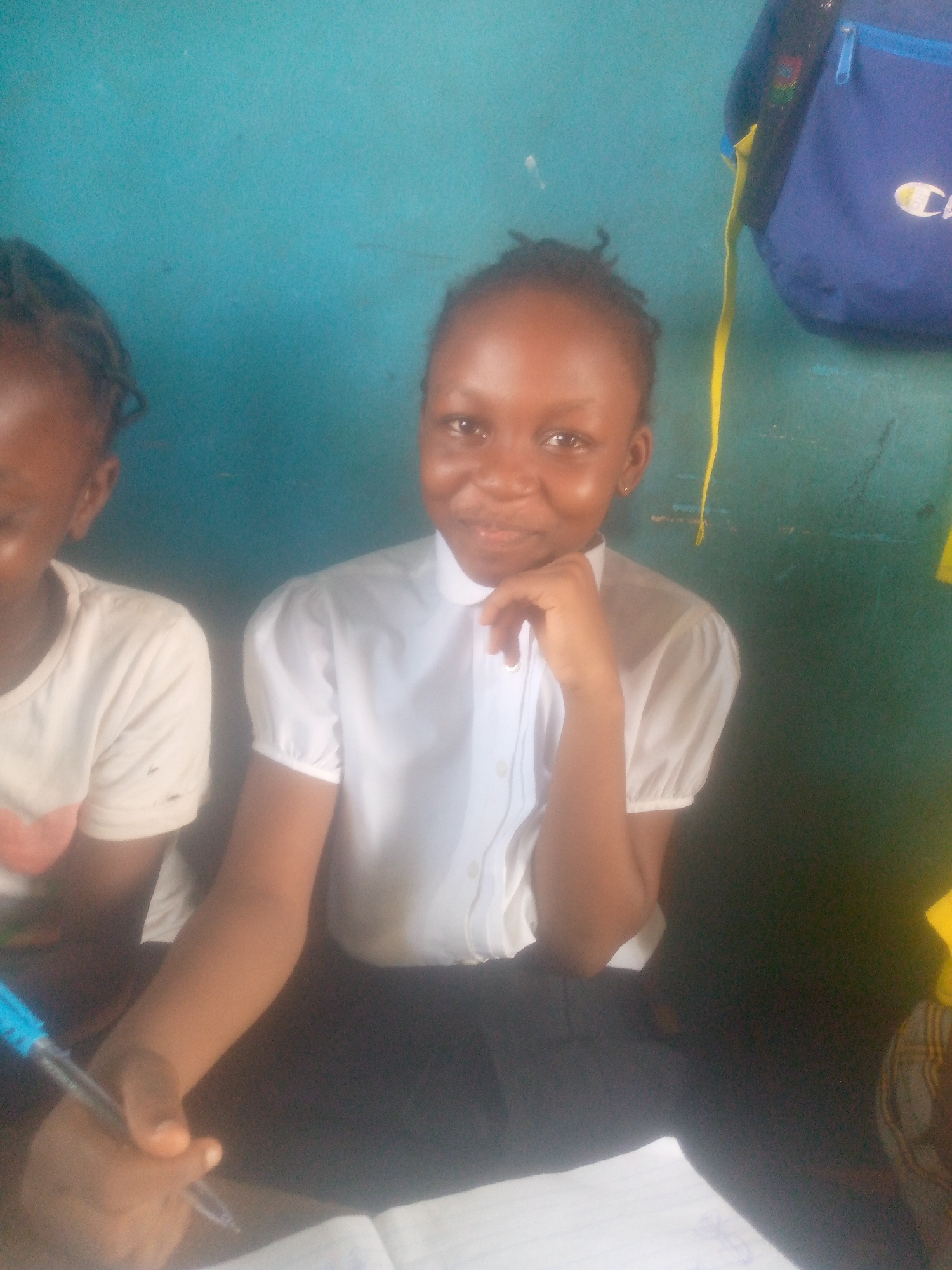The image depicts a young girl, likely a student, seated at a desk or table. She wears a white collared shirt and dark pants, possibly a school uniform. She is holding a pen and appears to be writing in a notebook. Her left hand rests on her chin. Another young girl is partially visible to her left, wearing a light-colored t-shirt. A blue backpack hangs in the background on a blue wall. The location appears to be an indoor setting, potentially a classroom, though the specific city and location are unknown.