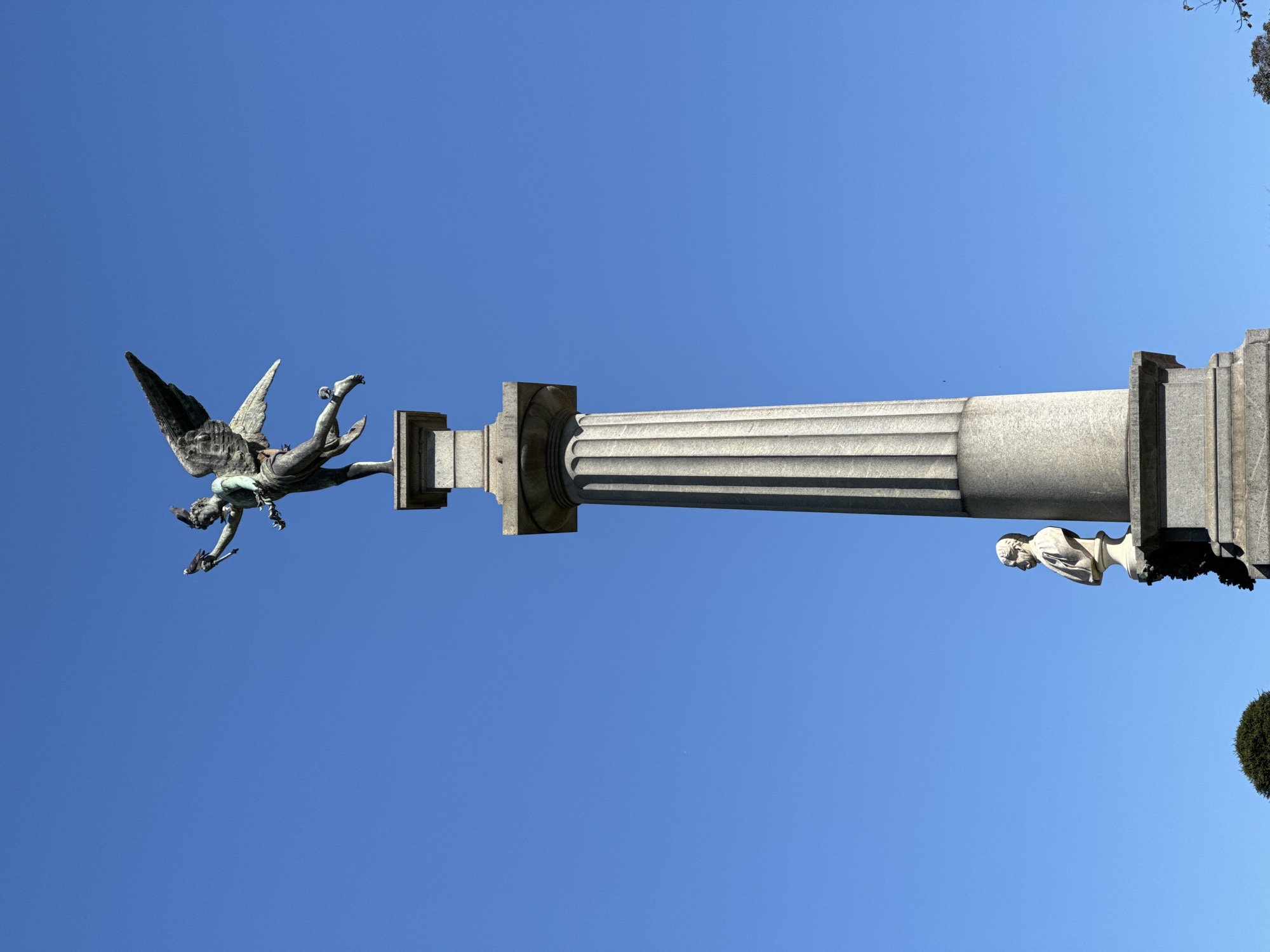 A horizontal, eye-level shot of the Recoleta Cemetery in Buenos Aires, Argentina, shows a stone pillar with a winged statue on one side and a bust on the other against a clear blue sky. The pillar is gray, fluted, and capped with square-shaped supports on each end. On the left end, a gray statue of a winged figure appears to be falling or flying downward, clutching a small object in its hand. On the right end, a white stone bust is perched on the pillar, facing away from the viewer. Green foliage is visible in the lower right corner, possibly from nearby trees or shrubs.