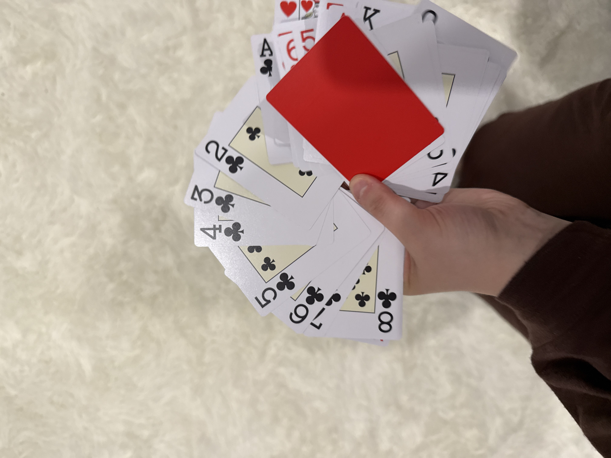 A hand holds a fan of playing cards, mostly of the suit of clubs, ranging from two to eight, along with an Ace of Clubs, five of hearts, King of hearts, and a blank red card. The background is a fuzzy, light-colored surface. A sleeve of a brown shirt is visible on the right edge of the image. The location is not discernible.