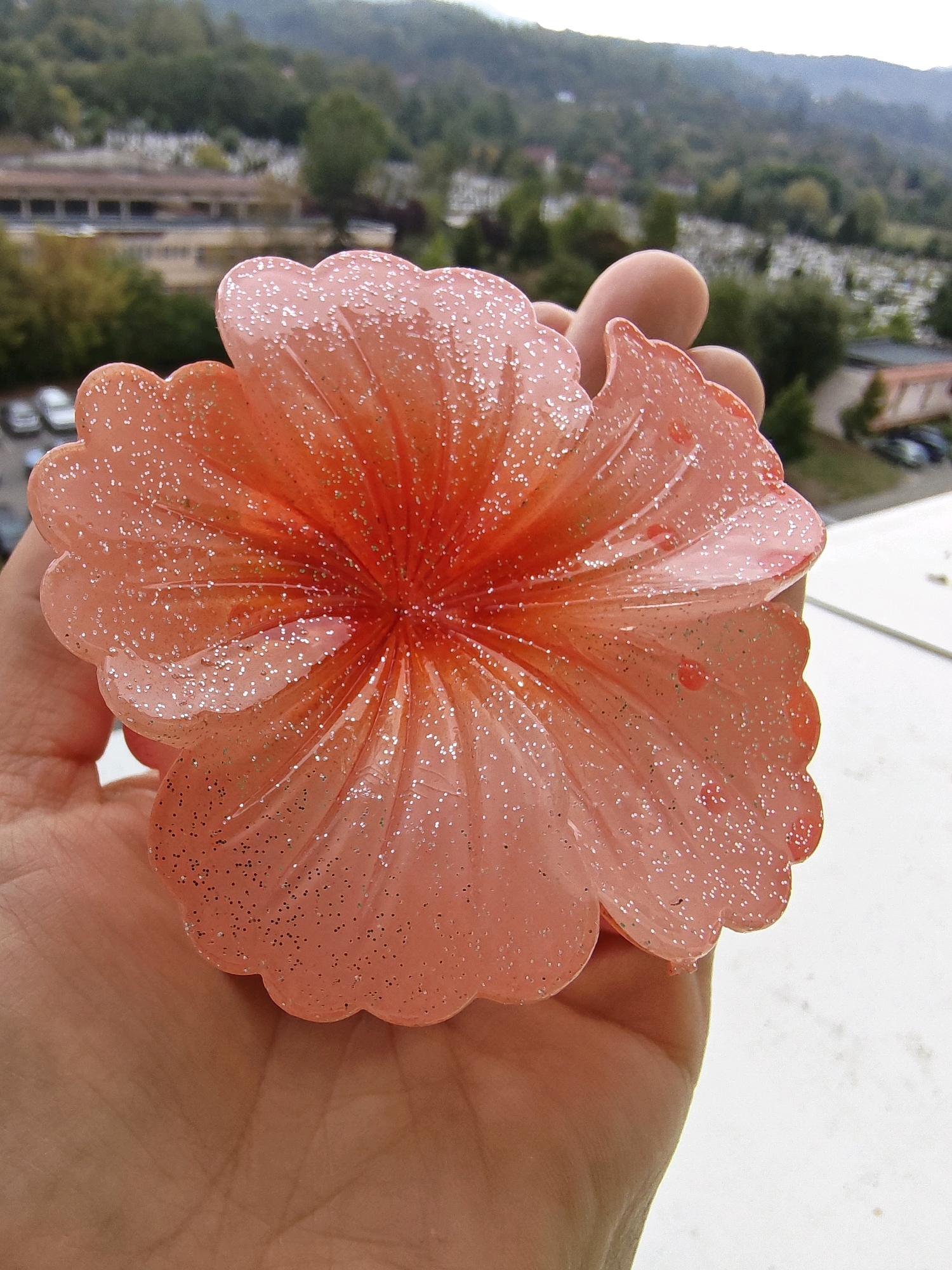 In Petroșani, Romania, a person's hand holds a translucent, peach-colored, glitter-infused decorative flower with a scalloped edge and a gradient of orange towards the center. The background is a blurred vista of a city with buildings, trees, and a faint outline of hills under an overcast sky.