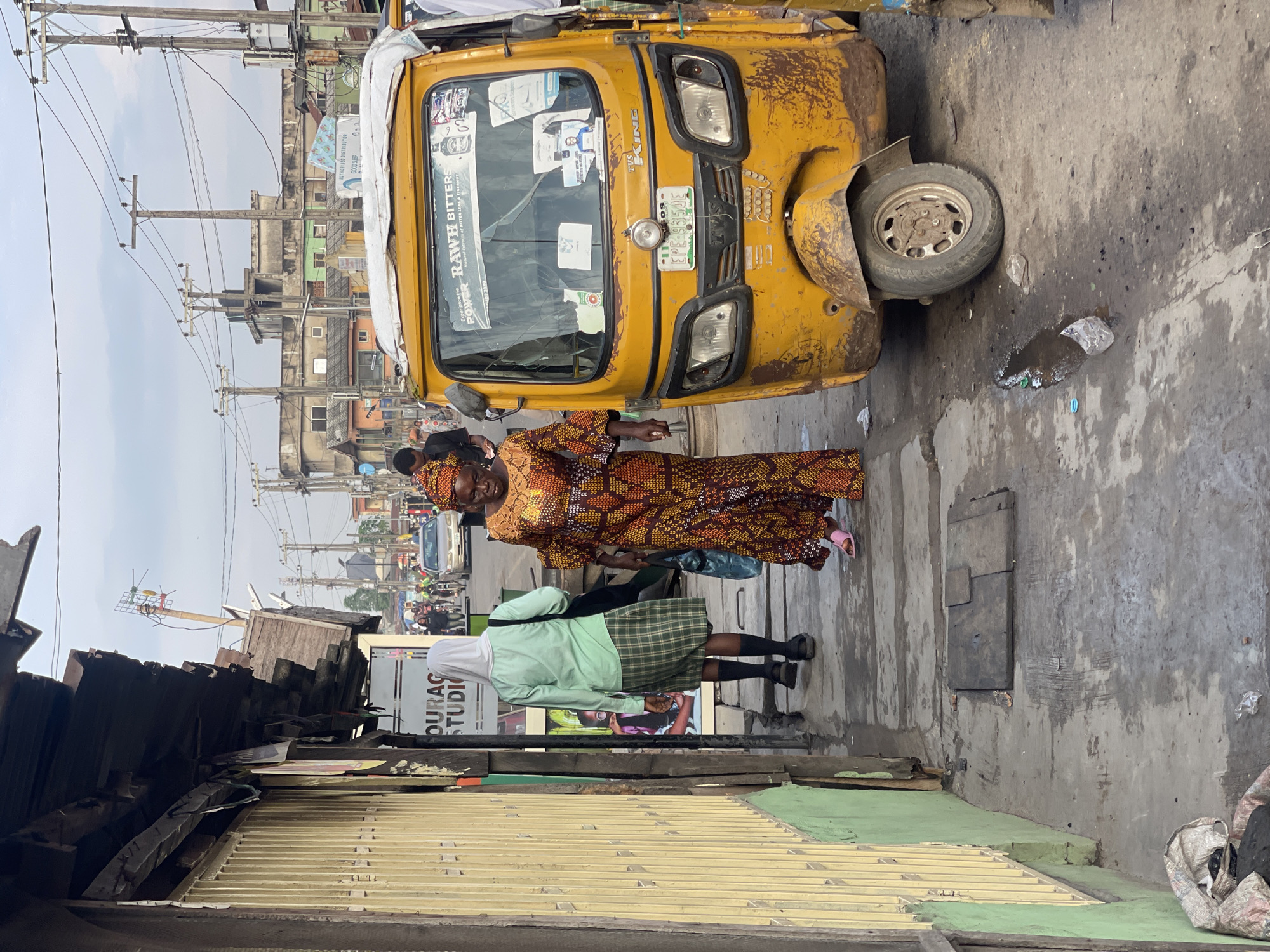 In Akowonjo, Nigeria, a yellow auto-rickshaw is parked on a street next to a sidewalk. The auto-rickshaw has a license plate that reads "EPE 933 JE". Two women are walking on the sidewalk. One woman is wearing a green shirt and a plaid skirt, and the other is wearing a colorful patterned dress. Behind them, a street scene unfolds with other people, vehicles, and buildings. The street is paved and appears wet in places. The buildings are a mix of concrete structures and corrugated metal, with visible electrical wires and poles overhead. The overall environment suggests a bustling urban area.