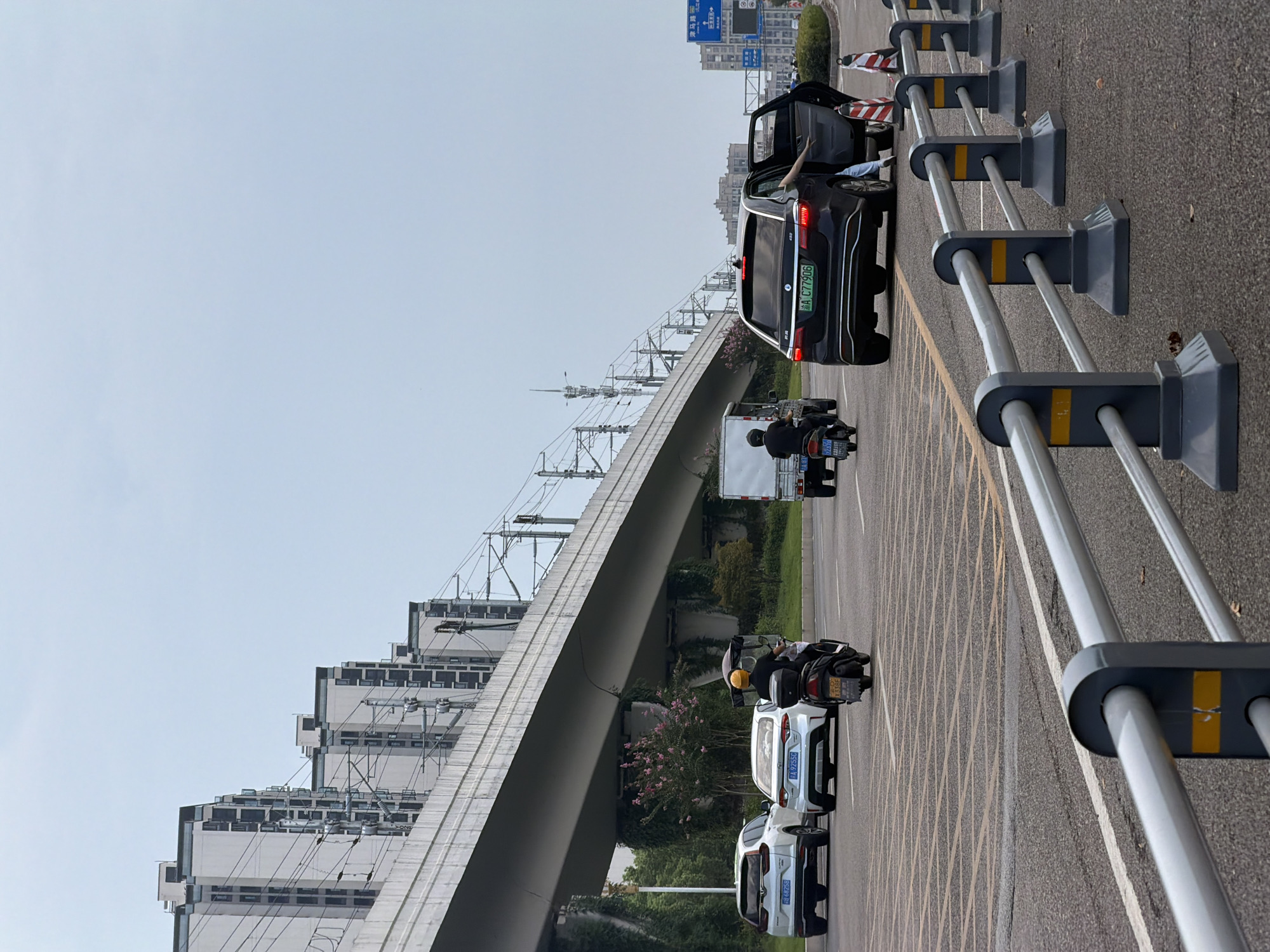 The image captures a street scene in Jiangjin District, Chongqing, China, under an overcast sky. An elevated railway or highway overpass dominates the upper portion of the frame, with numerous apartment buildings lining the distant embankment. In the foreground, a road features several vehicles. A black electric car with its driver's side door open is parked on the right, and a person is partially visible inside the car. Further left, a white car and a silver SUV are also parked on the roadside. A person on a black motorcycle with a yellow helmet is seen riding away from the viewer, and a white truck is visible in the middle distance. Traffic cones and a metal barrier line the edge of the road in the foreground. The overall environment suggests a city setting with ongoing urban development and transportation infrastructure.