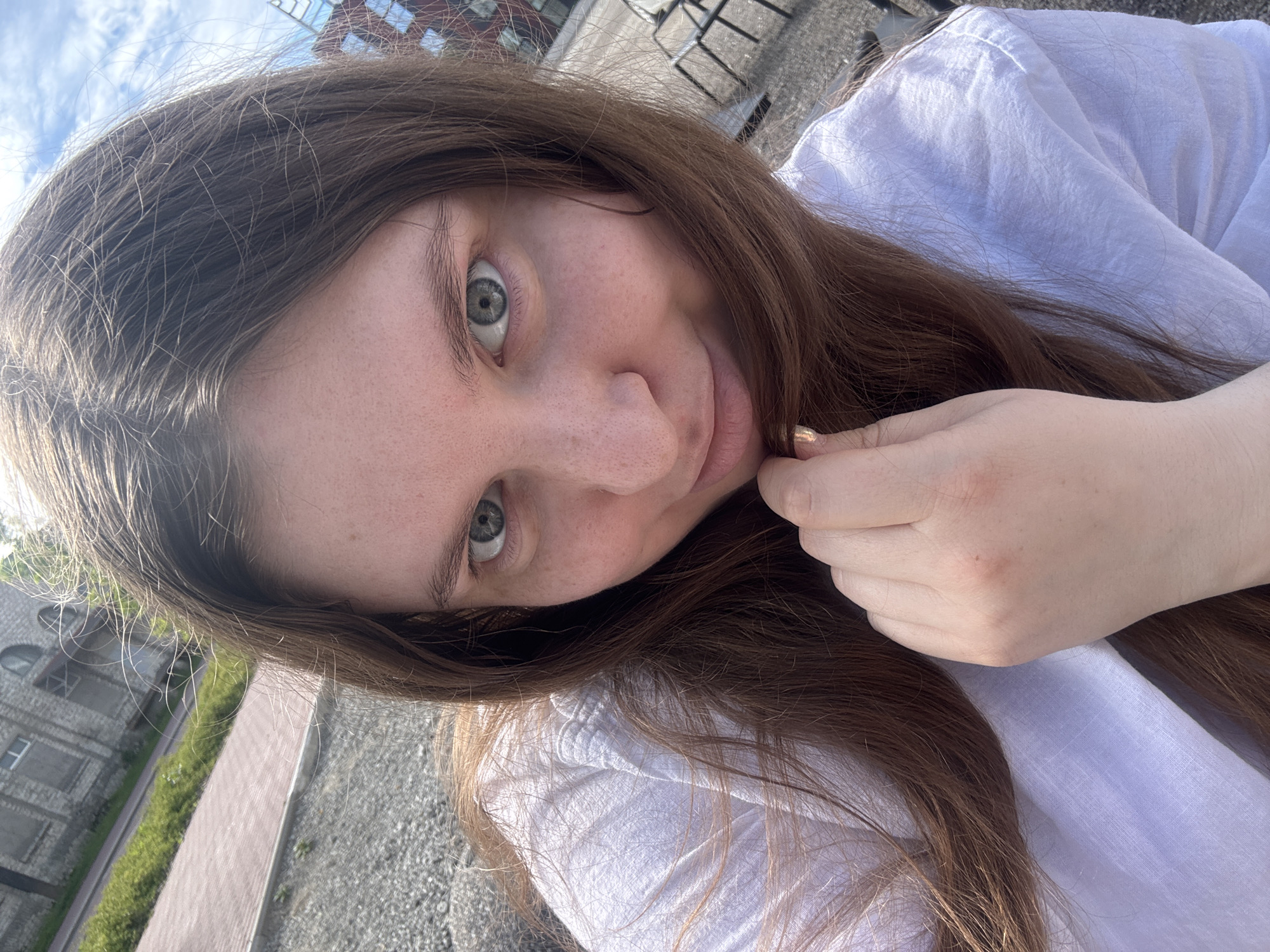 An outdoor, close-up, eye-level selfie shows a person with long, brown hair and light skin, with freckles. The person is wearing a white shirt. The person's right hand is visible, holding some hair near their chin. The background suggests an urban environment with a building that appears to be made of brick and concrete, with windows. There are also gravel and a paved walkway visible. The sky is partly cloudy. The scene is likely in Tallinn, Estonia.