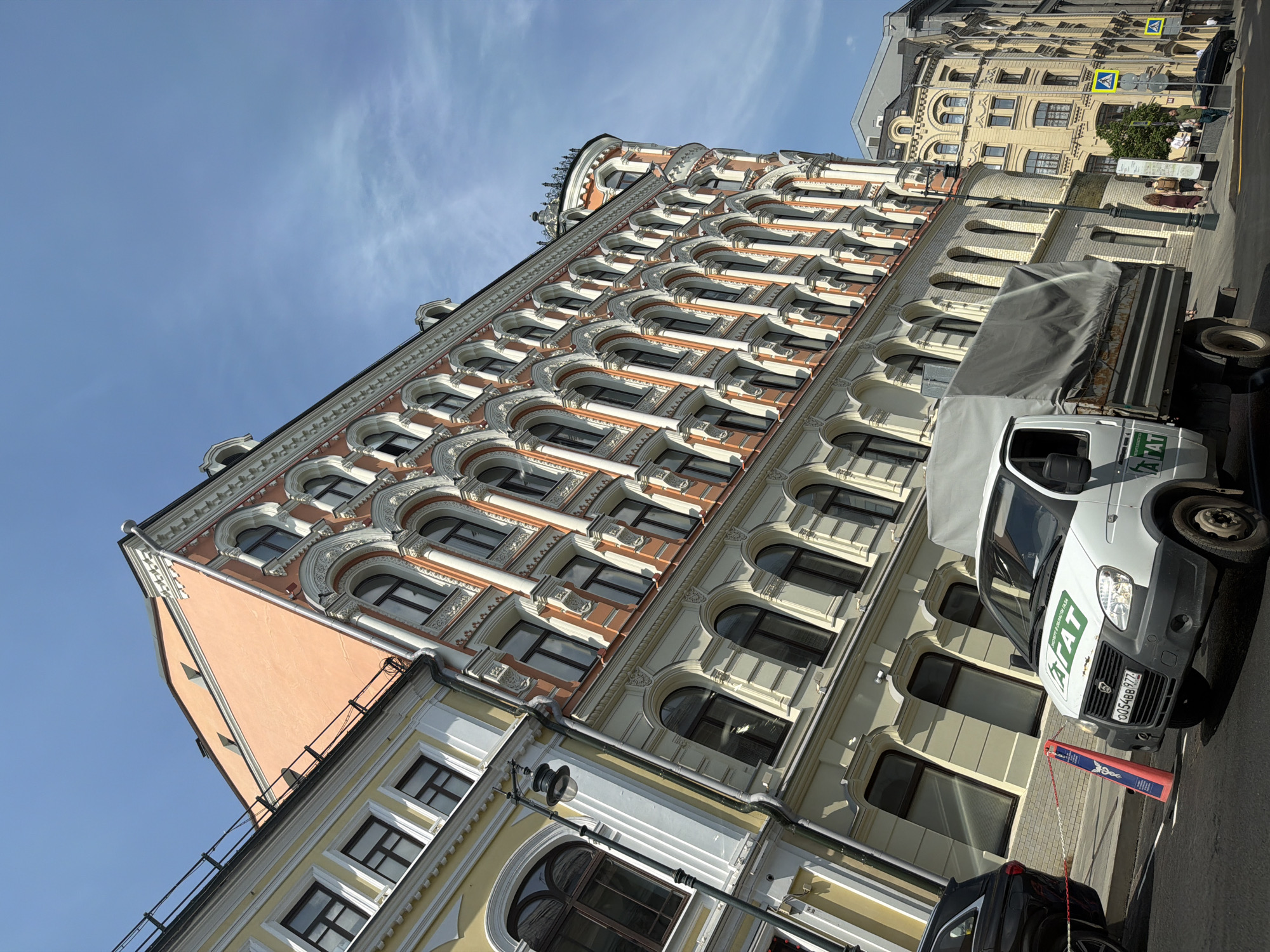 The image shows a street scene in Moscow, Russia. The camera points upwards at an angle, focusing on a set of buildings with ornate architectural details. The main building is a multi-story structure with arched windows and a peach-colored facade. The architecture is characterized by decorative molding and intricate design. Adjacent to it is another building with a yellow facade.
On the street level, there are vehicles, including a white van with the word "TAT" in green lettering, parked along the curb. A black car is partially visible in the foreground. Pedestrians are walking on the sidewalk, with a yellow pedestrian crossing sign visible in the distance.
The sky is blue with some scattered clouds.