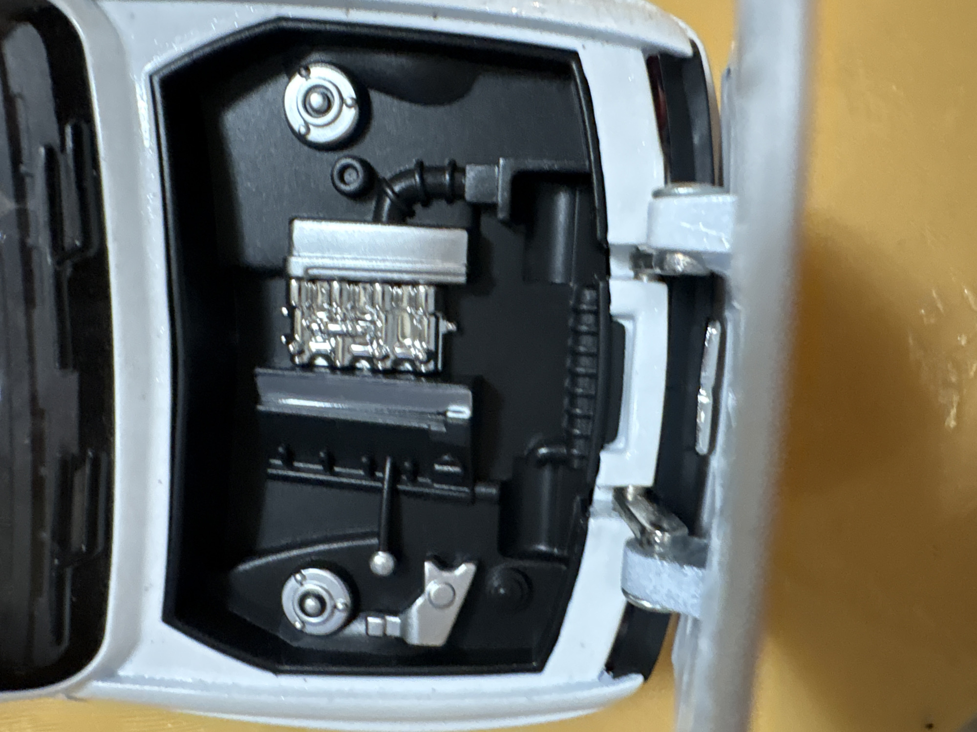 An overhead medium shot captures the highly detailed interior of a white toy car's engine bay, which is open. The engine itself is a miniature replica with intricate silver metal parts, including a silver manifold and what appear to be silver cylinders. Black hoses and black plastic components are also visible, mimicking a real engine. On the left side of the engine bay, a silver metal lever-like object is attached to the wall, and above it, a silver circular cap with screw-like details is fixed to the black surface. To the right of the engine, another similar silver circular cap is present. The background is predominantly black, representing the car's chassis. The hood of the car is open and is white on the outside and black on the inside. The bottom portion of the image shows a small part of the car's front bumper and hinges, with a yellowish surface beneath. There are no people in the image. The scene is indoors, likely in a controlled environment.
