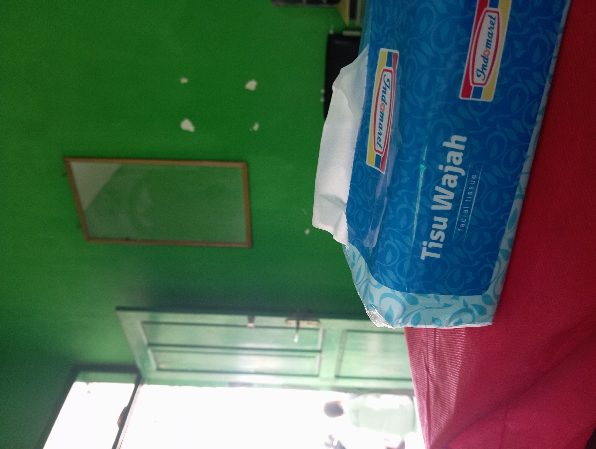 A packet of blue facial tissues, featuring the "Indomaret" brand and text in Indonesian reading "Tisu Wajah" and "facial tissue," is placed on a red surface. The packet has a floral pattern in a lighter shade of blue. In the background, a green wall is visible, with a framed picture hanging on it and a light-colored door with vertical panels and a dark handle. To the left, a bright light emanates from an open doorway, where a person wearing a white garment can be seen from behind. The overall scene suggests an indoor setting in a building, potentially a shop or residence, in Tegalciut, Indonesia.
