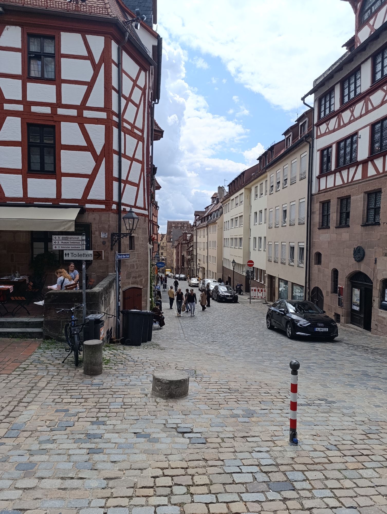 The image depicts a street scene in Nuremberg, Germany, likely in the historic old town, characterized by traditional half-timbered architecture. In the foreground, a cobblestone street leads uphill, lined with buildings. The building on the left features distinctive red and white half-timbering and a small outdoor cafe area with tables and chairs. A signpost points towards "Toiletten," "Zeugmuseum," "Museum Fembohaus," and "Historischer Kunstbunker." A bicycle is parked near the building.
Further up the street, the buildings continue, showcasing a mix of historical facades with intricate window designs and more modern apartment buildings with shutters. Several cars, including a black Audi, are parked or driving on the street. Pedestrians are walking along the sidewalk. The sky is partly cloudy.
A red and white striped bollard stands in the middle of the cobblestone street in the foreground. Several large black trash bins are visible near the corner of the building on the left. The overall impression is that of a picturesque, well-maintained historical district on a pleasant day.
