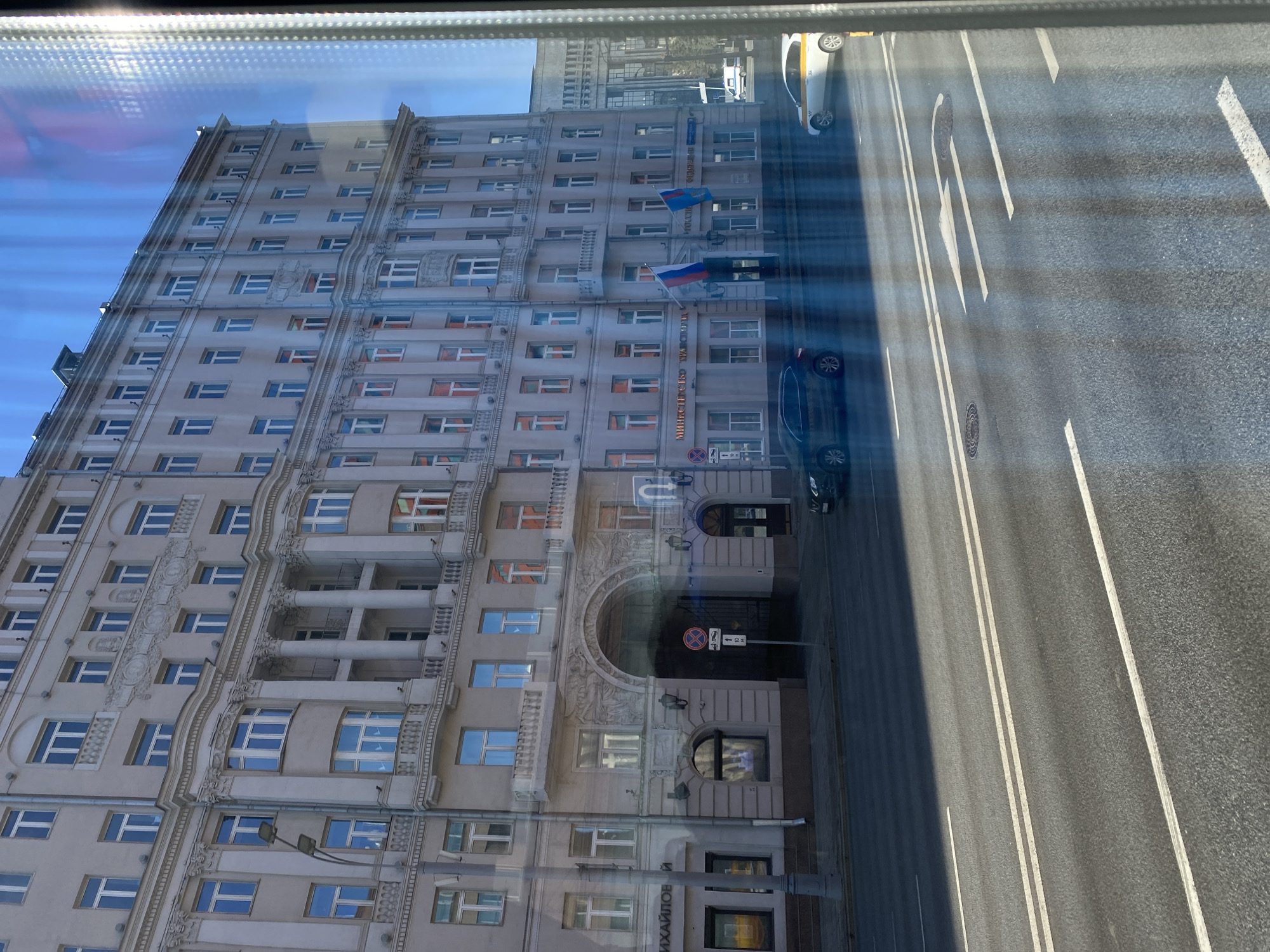 The image is taken from a window overlooking a street in Moscow, Russia. The building across the street is the Ministry of Foreign Affairs of Russia, identifiable by the flags displayed. A street with several lanes and road markings runs along the building. Cars are present on the road. The lighting indicates it is daytime. Reflections are visible on the window from which the picture is taken.