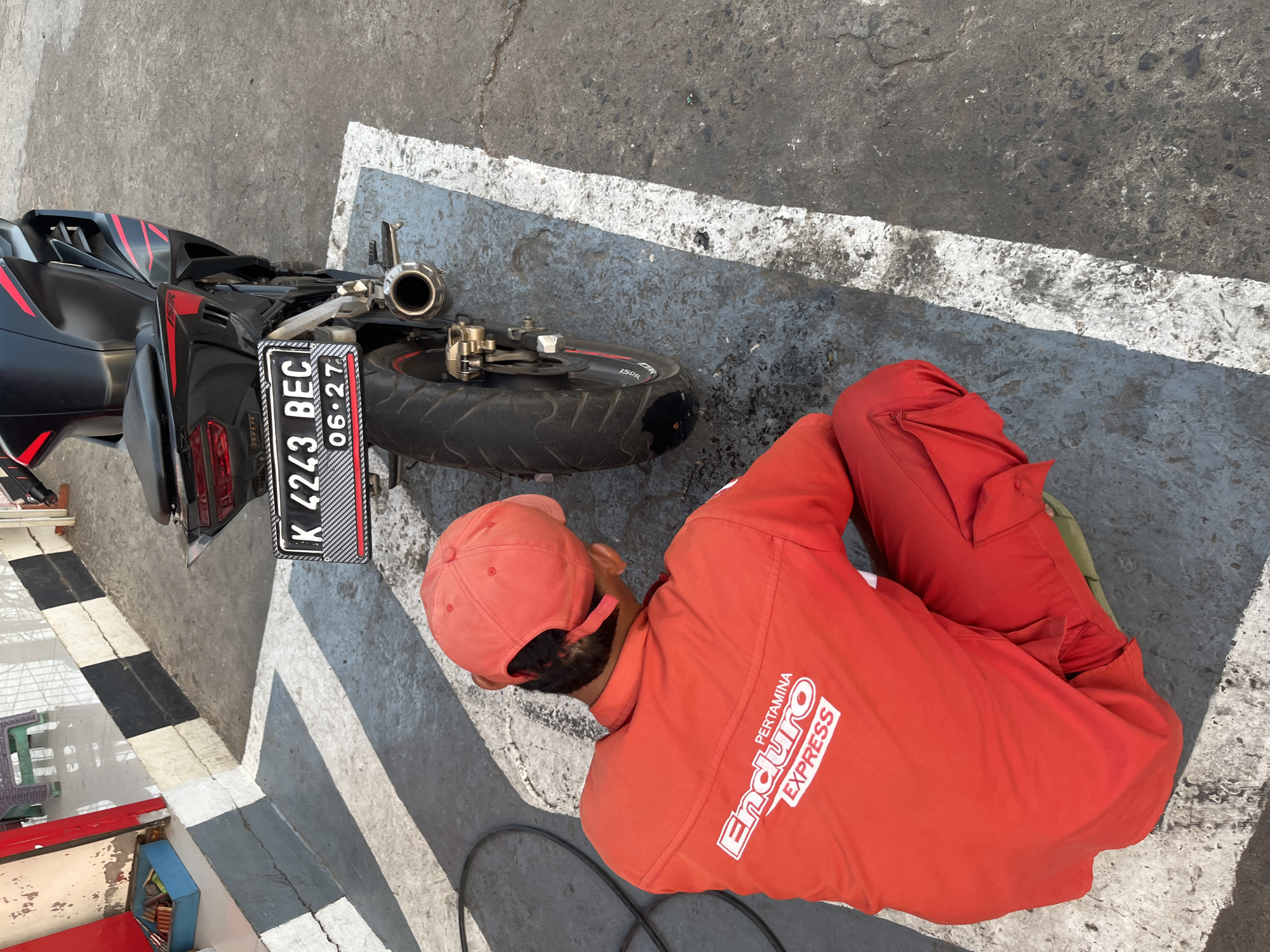 In Pecangaan, Indonesia, a man wearing a bright red uniform with "Enduro PERTAMINA EXPRESS" emblazoned on the back is kneeling on a gray concrete surface. He is facing away from the camera, towards a black motorcycle with red accents parked on a white-lined section of the ground. The motorcycle has a license plate that reads "K 4243 BEC" and "06-27". The environment appears to be an outdoor area, possibly a service station or parking lot, with black and white striped markings on the ground and a portion of a red building visible in the background. The lighting suggests it is daytime, casting shadows on the ground.