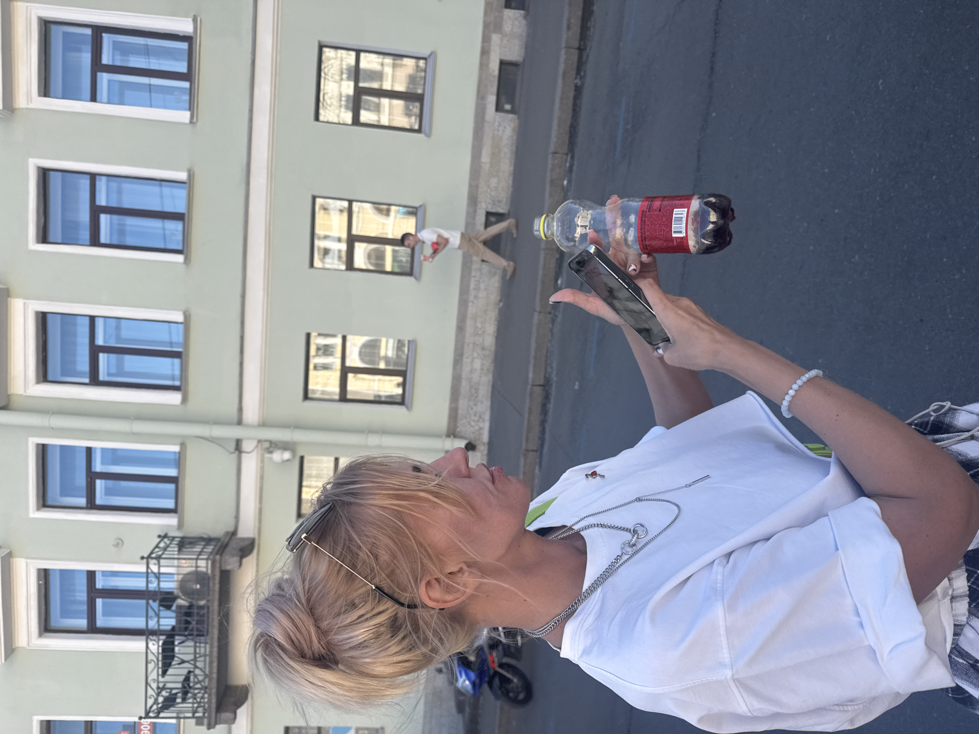 A young Caucasian woman with blonde hair pulled into a high ponytail stands on a city street, holding a smartphone in her right hand and a partially filled plastic bottle of a dark beverage in her left. She is wearing a white t-shirt, a plaid skirt, and several silver necklaces. Behind her, a light green building with several windows and a small balcony is visible. In the background, a man in light-colored pants and a white shirt walks along the sidewalk, carrying a red object. A blue motorcycle is parked on the left side of the street. The scene is likely in Saint Petersburg, Russia, given the architectural style of the building.