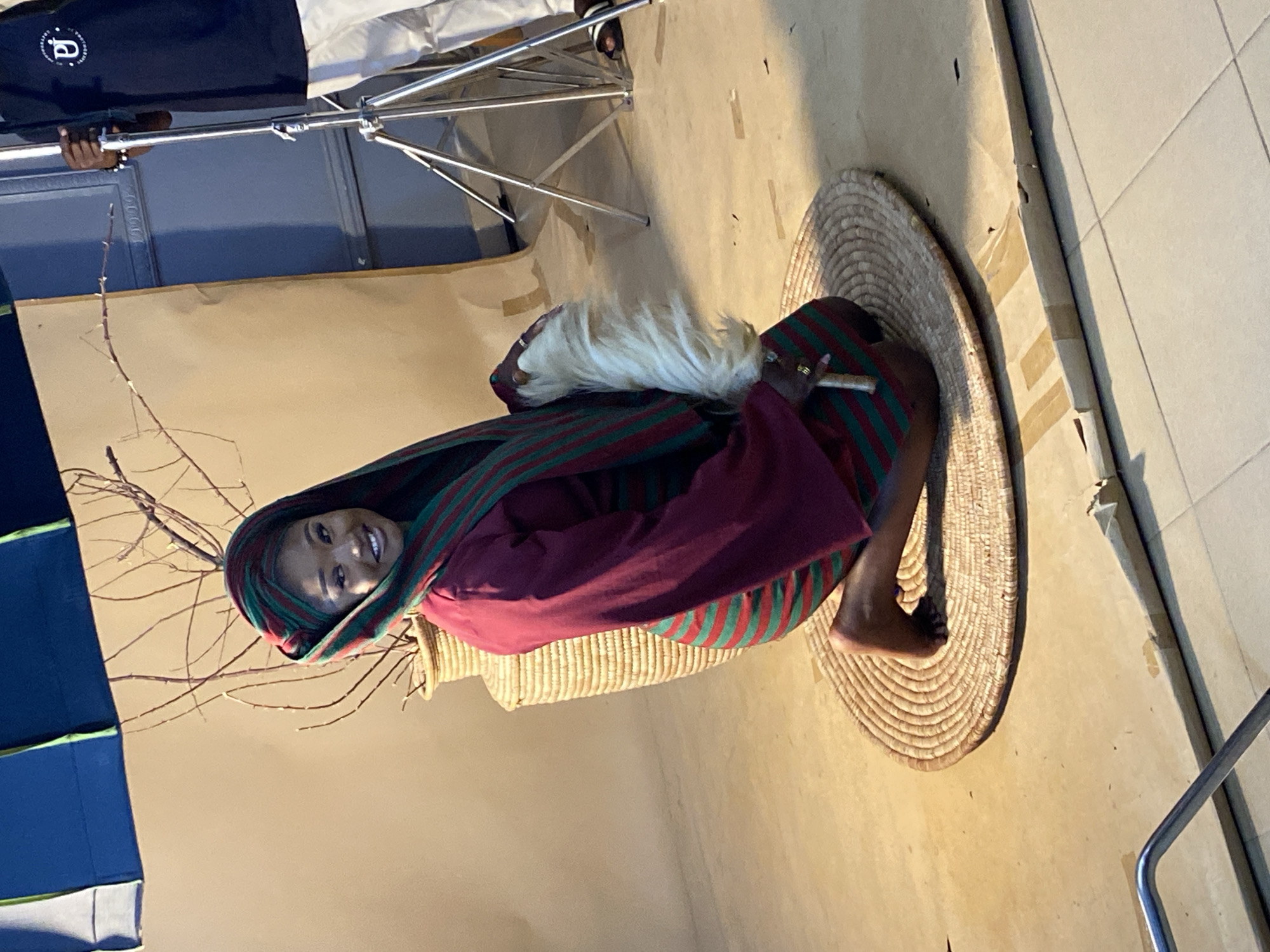 In an indoor studio setting in Bukuru, Nigeria, a young Black woman kneels on a circular woven mat. She is dressed in a maroon garment with green stripes and a matching headscarf. She holds a white feathery object in her right hand and wears rings on her fingers. Behind her, a large woven basket with dried branches stands against a plain backdrop. To the right, a photographer, partially visible, holds a light stand. The floor is a tiled surface partially covered with large pieces of cardboard. The overall scene suggests a professional photoshoot.