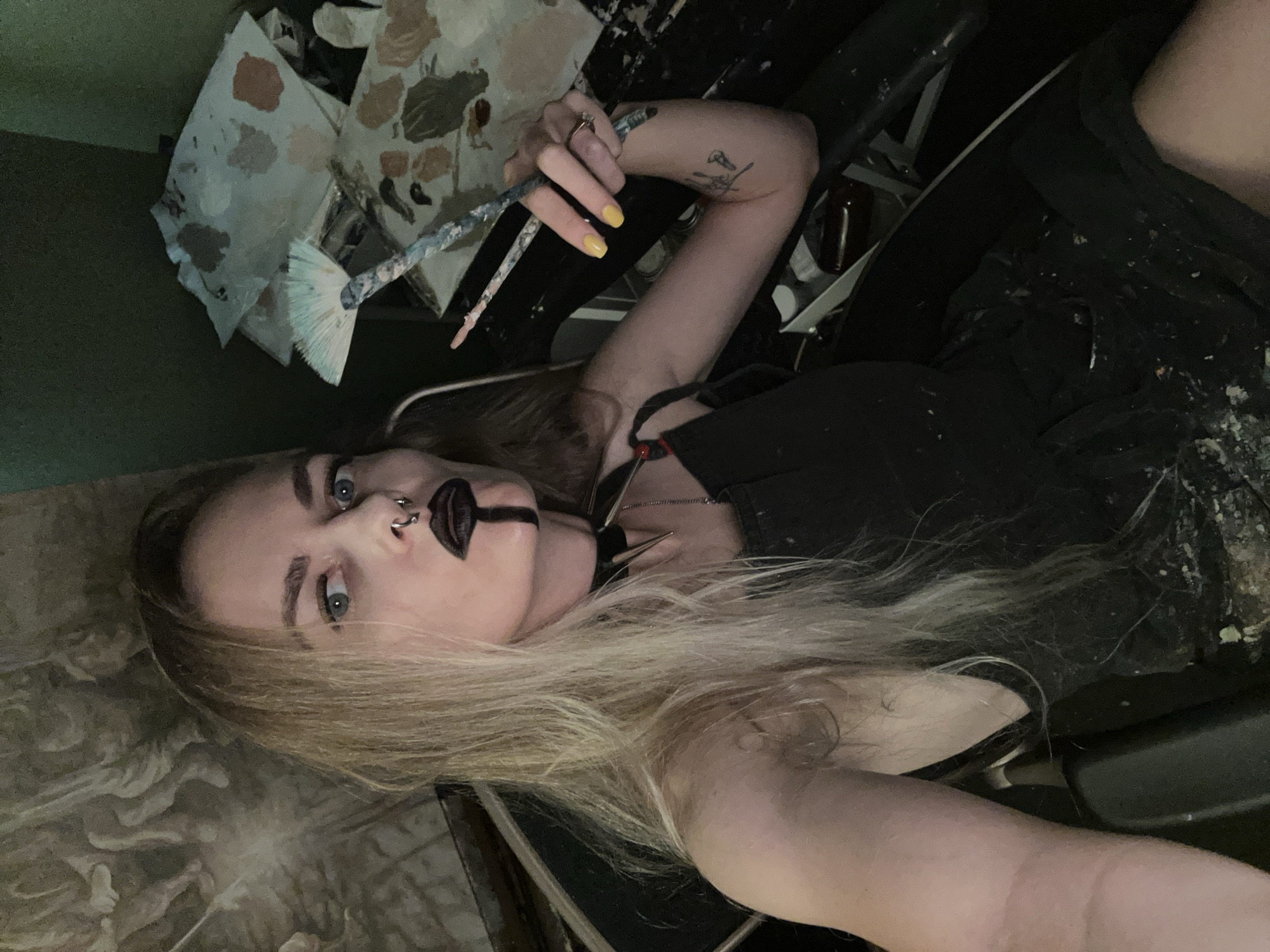 A blonde woman with blue eyes and a nose ring, wearing black lipstick and a spiked choker, sits in a black chair holding paintbrushes. She is wearing a black apron that is splattered with paint. In the background, a large mural or tapestry is visible on the wall. There are several paint palettes with various colors of paint on them. The scene appears to be an artist's studio, possibly located in Cape Coral, Florida, United States of America.