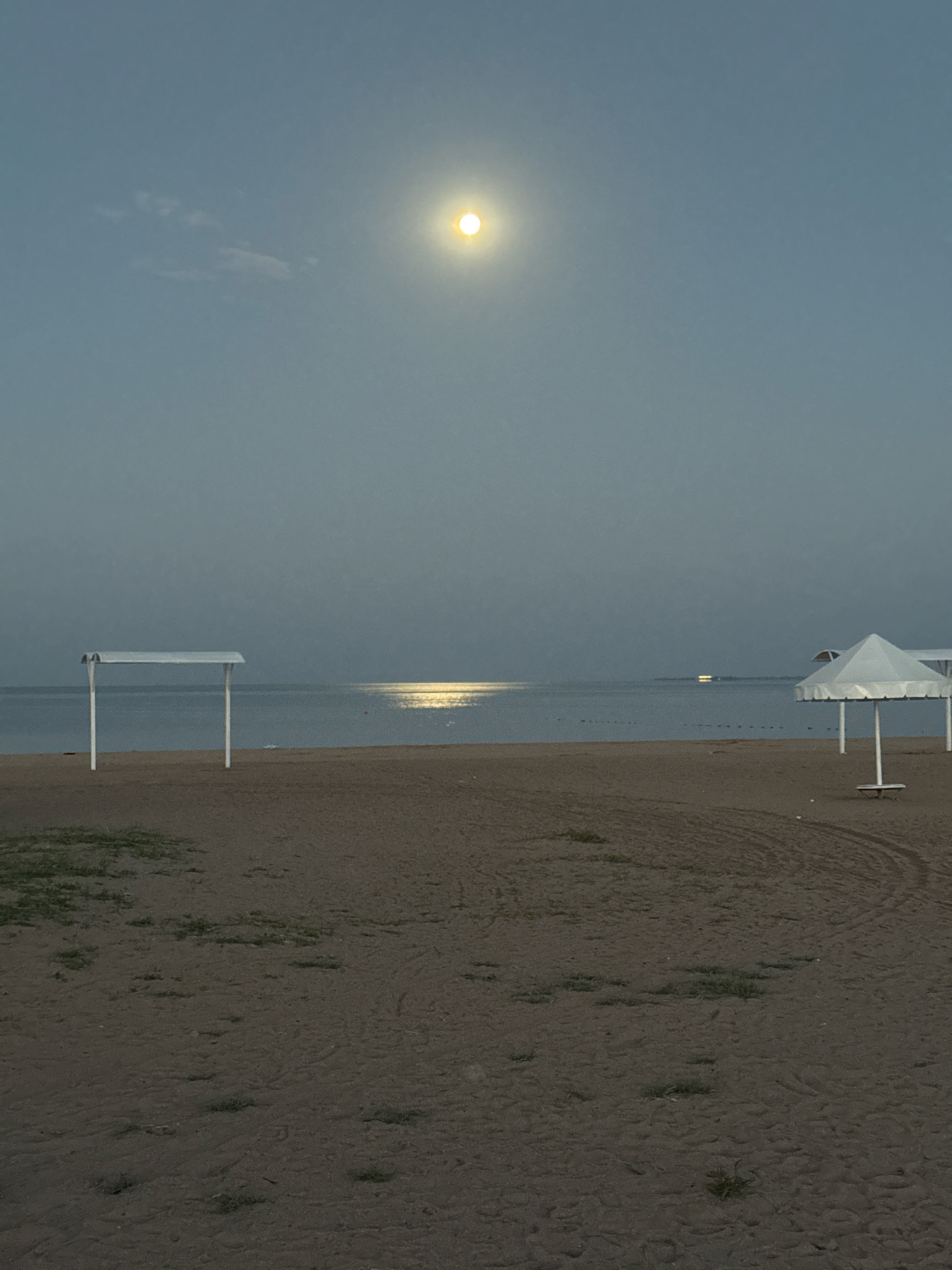 The image depicts a serene beach scene in Bosteri, Kyrgyzstan, likely during the evening or early morning, with the moon prominently visible in the sky. The moon, a bright, yellow orb, is positioned in the upper center of the frame, casting a shimmering reflection on the calm water. The beach itself is sandy, with tire tracks visible in the foreground, suggesting recent activity. Two white structures, possibly beach umbrellas or shelters, are present on the sand; one is on the left side of the frame and another on the right. The water stretches out to the horizon, where faint lights are visible in the distance, possibly from boats or a settlement. The overall atmosphere is tranquil and peaceful.