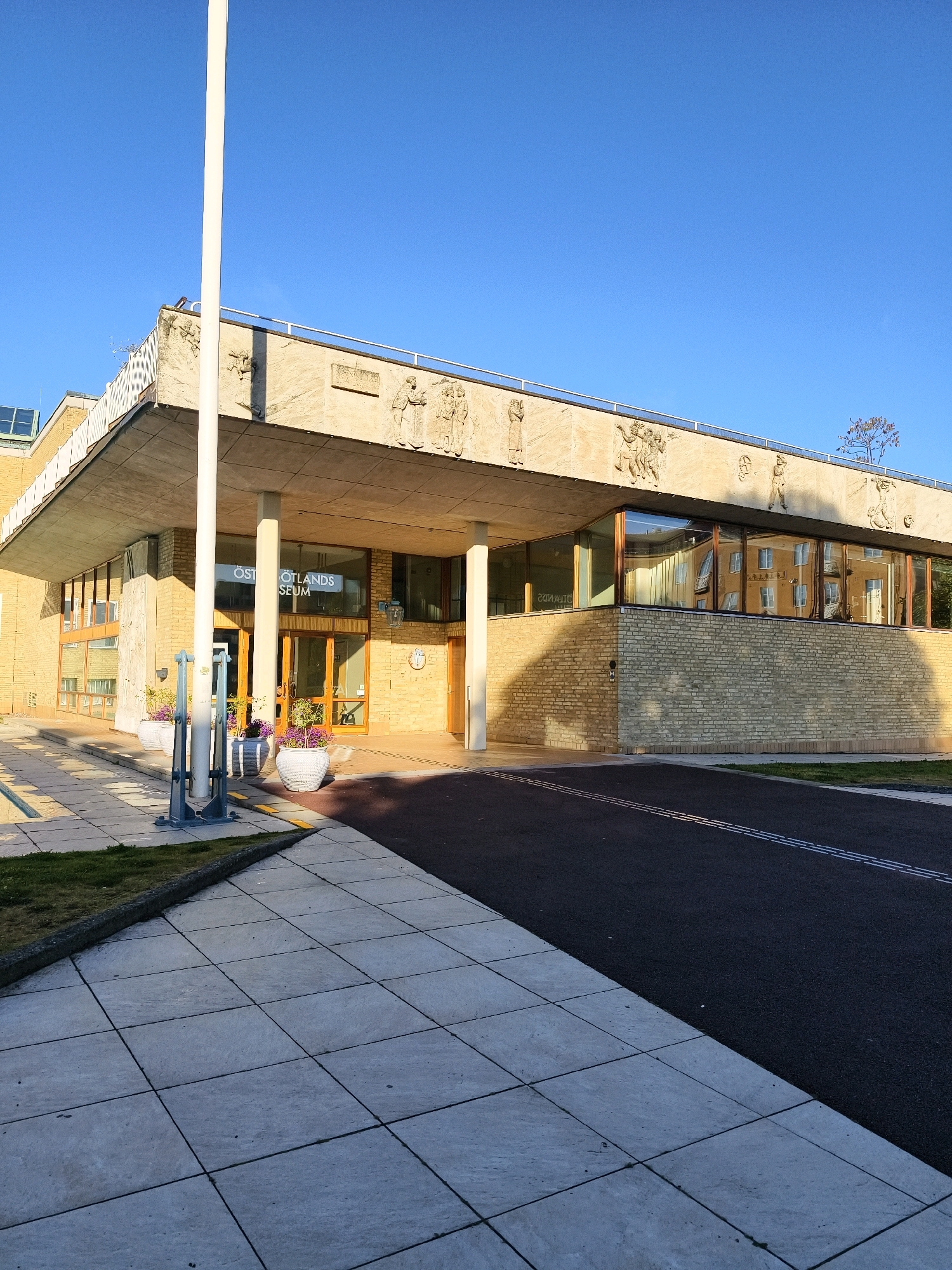 The image shows the Östergötlands Museum in Linköping, Sweden, under a clear blue sky. The building features a modern architectural style with a large concrete overhang supported by columns. The facade is primarily constructed of light-colored brick, and the upper section of the building has stone relief carvings. Large windows reflect the surrounding environment, including nearby buildings and trees. In front of the entrance, there are planters with purple flowers and a tall white flagpole. The foreground is a paved walkway made of large square tiles, with a section of asphalt road running alongside it, marked with double white lines. The scene is bathed in the warm light of what appears to be either early morning or late afternoon. There are no people visible in the image.