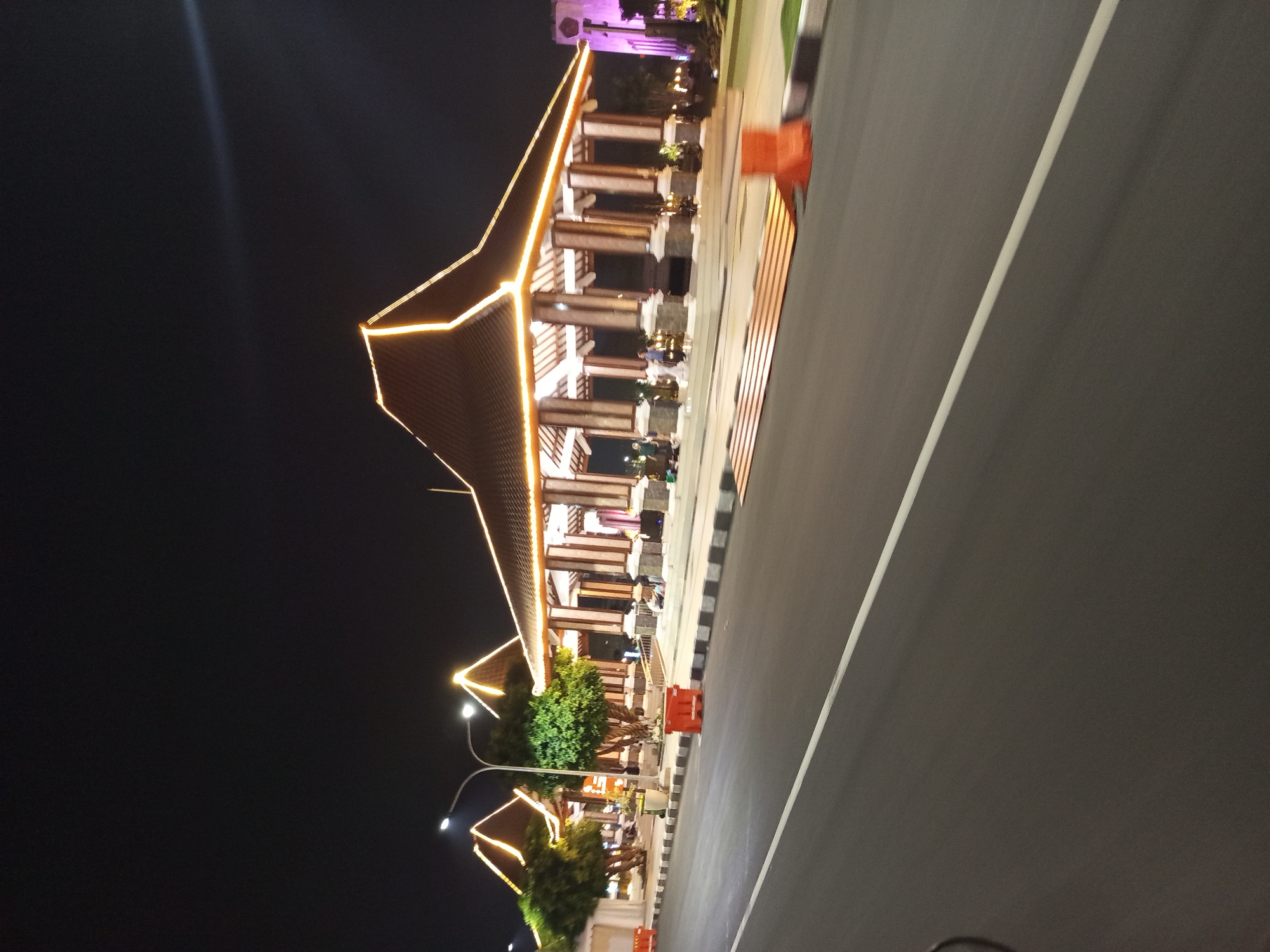 The image depicts a night scene in Sidoarjo, Indonesia, featuring a traditional Indonesian pavilion illuminated by string lights. The pavilion, with its distinctive layered roof, is supported by numerous columns. The area in front of the pavilion is paved and lined with barriers. A road with a white lane marker runs in the foreground, suggesting a public space or a roadside attraction. Several trees are visible behind the pavilion. The lighting indicates it is nighttime, and the warm glow of the lights creates a festive atmosphere. There are a few individuals scattered around the pavilion, some appearing to be seated or walking. The overall environment appears to be clean and well-maintained.
