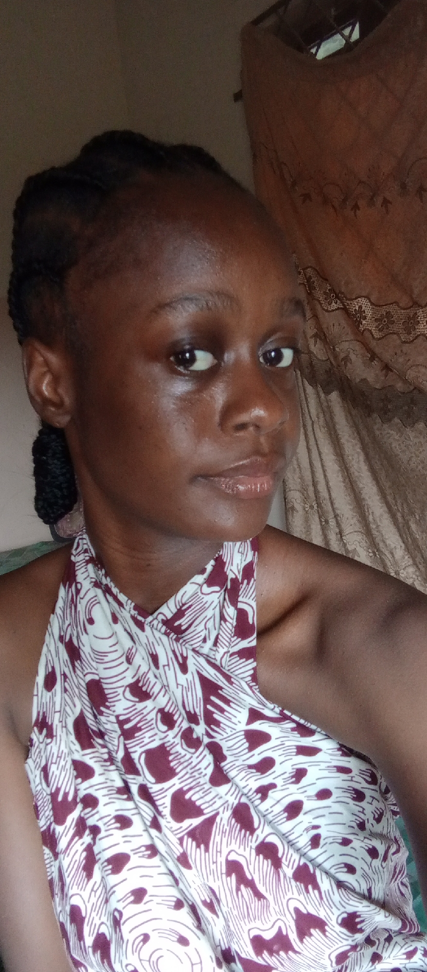 A young woman from Akwanga, Nigeria, is captured in a close-up, chest-up portrait. She has dark skin and her hair is styled in neat braids pulled back from her face, with a bun or coil visible behind her left ear. Her face appears naturally radiant, with a subtle sheen on her skin, and her lips have a light gloss. She looks directly at the camera with a calm, neutral expression.
She is wearing a halter-neck top that is light-colored, possibly white or off-white, and features a distinctive pattern of maroon or dark red abstract shapes, some resembling stylized hands or organic forms. The fabric of her top drapes softly around her neck and shoulders.
The setting appears to be indoors during the daytime, suggested by the natural light illuminating her face. The background on her left is a plain, light-colored wall. To her right, a textured, possibly brown or beige curtain with a decorative lace or floral pattern at the top is visible, suggesting a window or room divider. There is no visible text in the image.