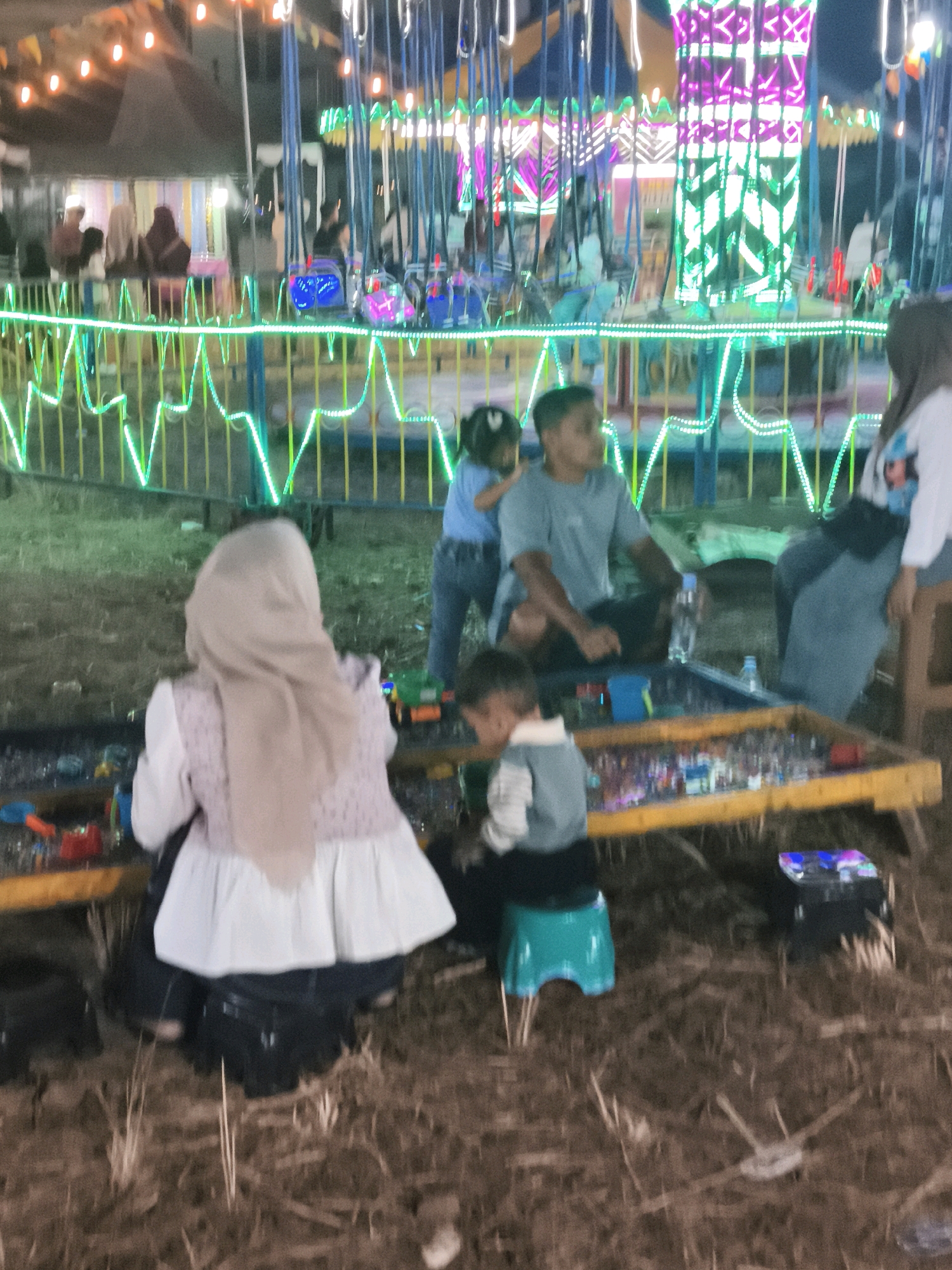 The image depicts an outdoor night scene, likely at a fairground, with a focus on several individuals and an amusement ride in the background. The ride features illuminated poles and hanging swings, with bright green and pink lights outlining its structure. In the foreground, a table filled with small toys or game prizes is set up.
Several people are visible. A woman in a light-colored hijab and a patterned vest over a white top is seated facing away from the camera, looking at the table. Next to her, a young boy in a light-colored shirt with a darker collar is also seated, appearing to interact with the items on the table. Further back, a man in a gray t-shirt is seated, and to his left, a young girl in a blue shirt is standing. Another woman, wearing a white top with colorful graphics and dark pants, is seated on a wooden stool on the right side of the frame, facing left.
The environment appears to be a sandy or dirt ground, with dry grass and straw scattered around. The overall impression is a lively, albeit slightly blurry, scene of nighttime entertainment. Given the context of fairs and amusement rides, this scene could potentially be located at a temporary event space within the city of Palembang, Indonesia, such as a public square or a designated fairground often set up for festivals or holidays.