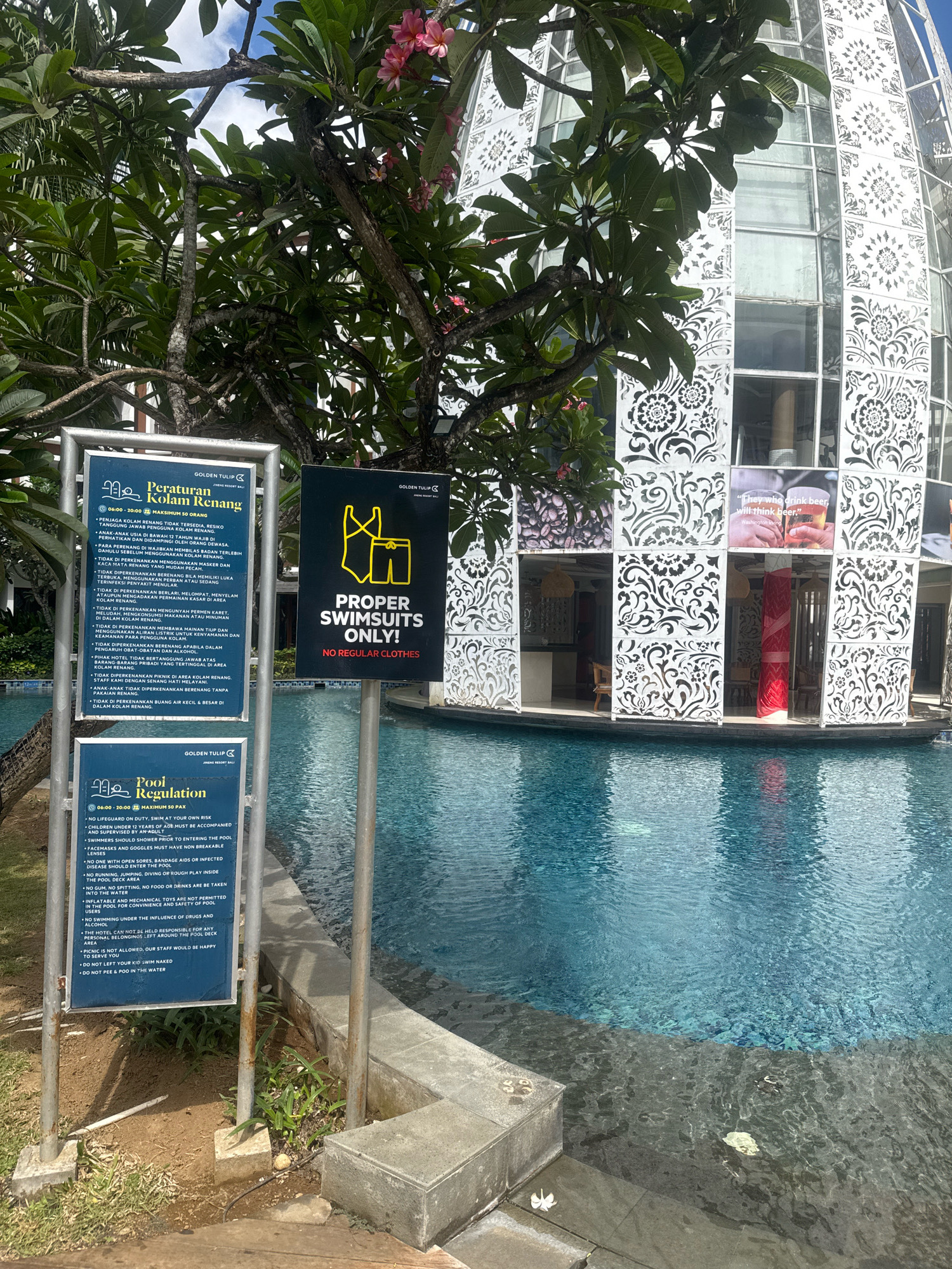 A bright, sunny day at an outdoor swimming pool in Kuta, Indonesia, likely at the Golden Tulip Jineng Resort Bali. The pool is a large, clear blue, with reflections of the sky and surrounding buildings on its surface. To the left, a lush green tree with pink flowers hangs over the water. In front of the tree, a metal signpost holds two blue signs with pool regulations. The larger sign on the left is written in Indonesian and has the title "Peraturan Kolam Renang" (Swimming Pool Regulations). The smaller sign below it is titled "Pool Regulation" and lists rules in English. To the right of the signs, a yellow sign with a graphic of swimwear and the text "PROPER SWIMSUITS ONLY! NO REGULAR CLOTHES" is mounted on a pole. In the background, a modern, circular building with decorative white patterns on its facade and large windows stands beside the pool. Some of the windows display advertisements or artwork. The overall scene depicts a well-maintained resort environment with clear rules for pool usage.