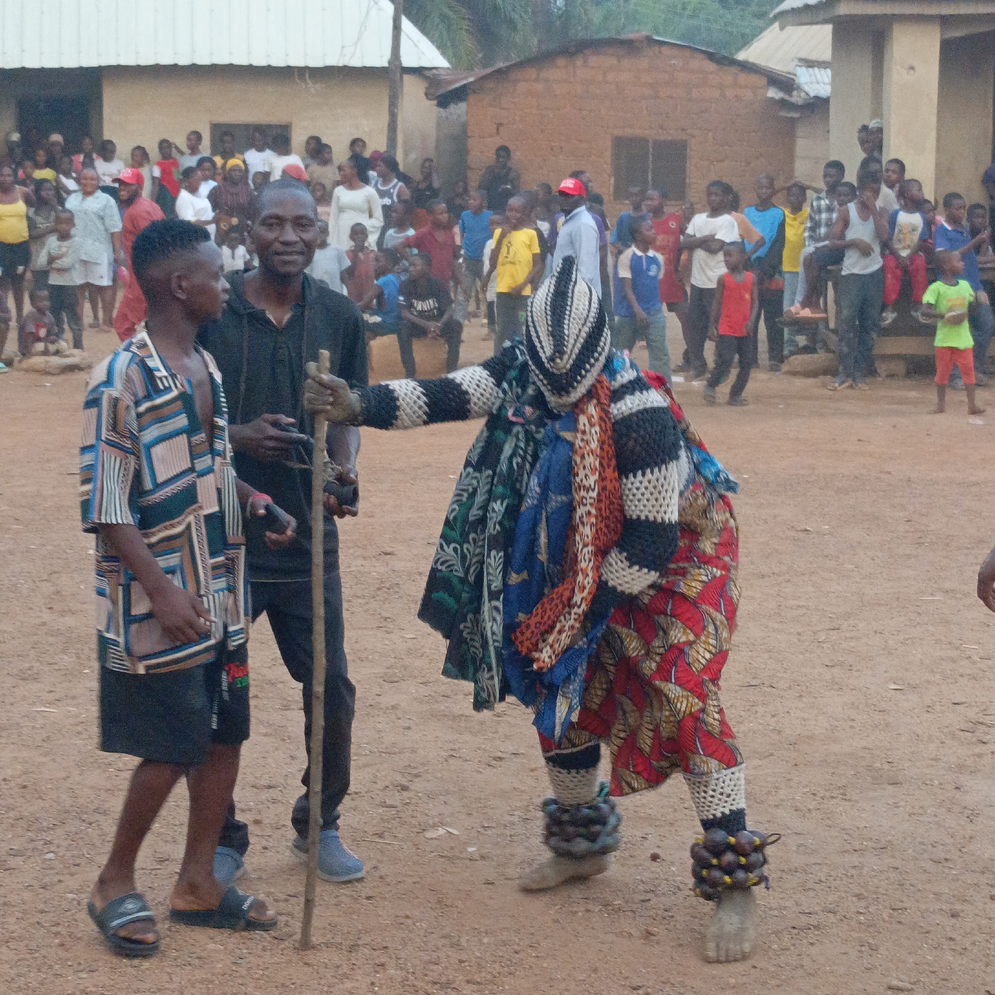 The image depicts an outdoor scene in Akwanga, Nigeria, where a crowd of people have gathered on a dirt ground surrounded by low buildings. In the foreground, there are three men: One man wears a short-sleeved geometric patterned shirt, black shorts, and sandals. Next to him, a man in a black hooded jacket and trousers holds a wooden stick. To their right stands an individual in a traditional masquerade costume, which includes a black and white knitted head covering, various patterned cloths draped over the body, a red and yellow patterned skirt, and leg coverings with attached round objects. The masquerade figure is gesturing towards the man in the black jacket with their outstretched hand. In the background, a large group of spectators watch the scene. The architecture consists of simple, one-story buildings with varying roof materials.