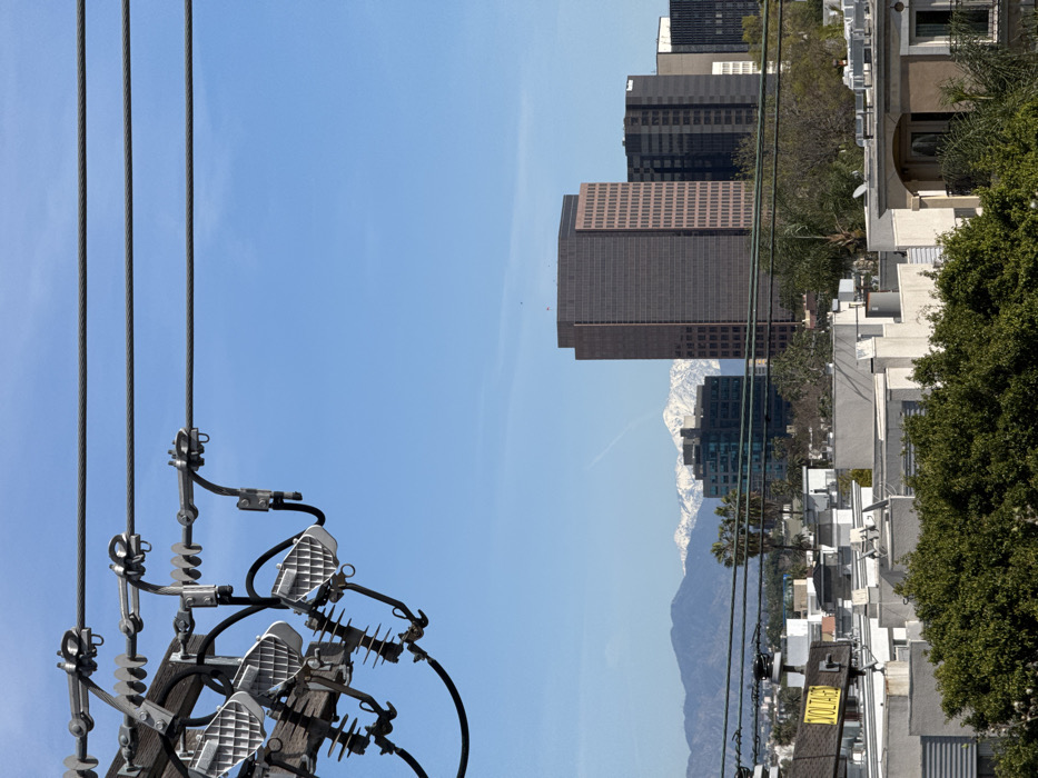 A cityscape view features a clear, blue sky. Power lines and utility poles dominate the left foreground. In the distance, tall buildings rise against the sky, with a mountain range visible beyond. Residential buildings and greenery are present on the right side, adding contrast to the urban scene. The image is rotated, causing the sky to appear on the left.