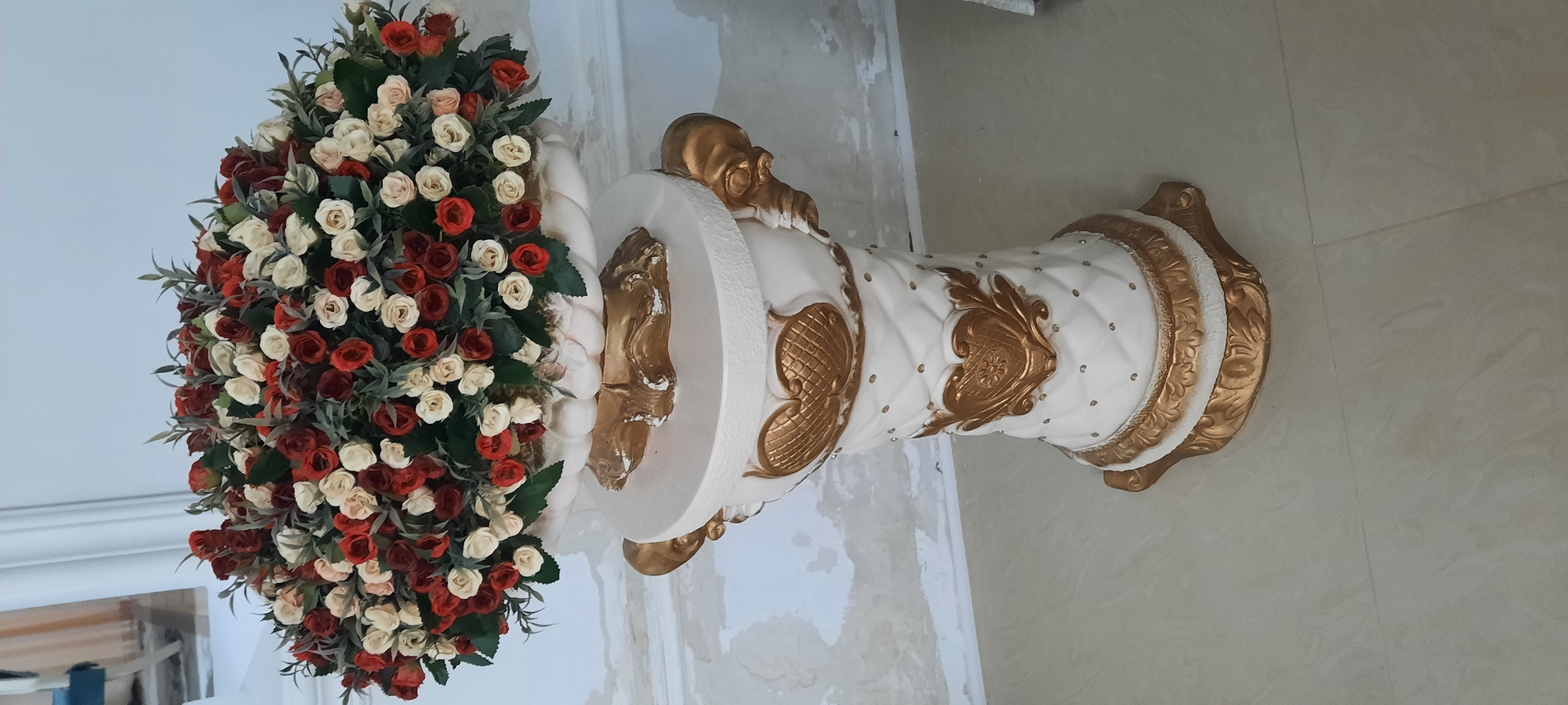The image displays a decorative arrangement consisting of a large bouquet of artificial flowers and a ornate white and gold pedestal. The bouquet, positioned on the left side of the frame, is densely packed with red, cream, and blush-colored roses interspersed with green foliage. The pedestal, on the right, is cylindrical with a flared base and top. Its surface is textured and features raised gold decorative elements, including swirling patterns and ornate crests. Small, clear embellishments are scattered across the white surface of the pedestal. The background appears to be an indoor setting, with a tiled floor and a wall that shows signs of wear or unfinished plaster. The lighting suggests an indoor environment with ambient light, and there are no clear indicators of time of day or weather. The image is oriented vertically, as if the camera was held upright. The overall impression is of a decorative display, possibly for an event or as a permanent fixture in a room. There is no visible text in the image. The context of Gudermes, Russia is provided but not visually evident within the image itself.