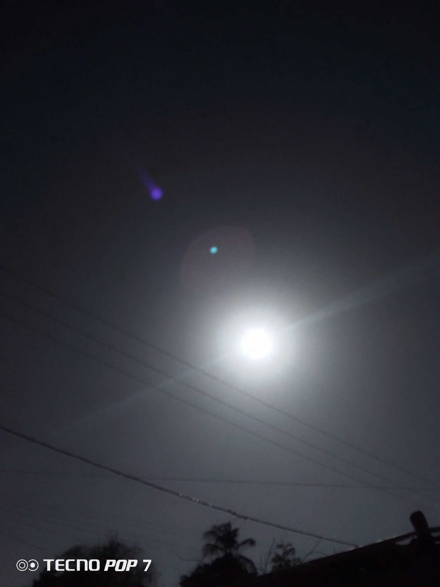 The image shows a bright light source, possibly the moon or a strong street light, in a dark sky. Power lines are visible silhouetted against the light. Below, there are outlines of trees and rooftops. The TECNO POP 7 watermark indicates the phone model used to capture the image. The location cannot be determined to be Altagracia, Venezuela, based on the image alone. There are no visible people, subjects, interactions, or explicit actions in the frame.