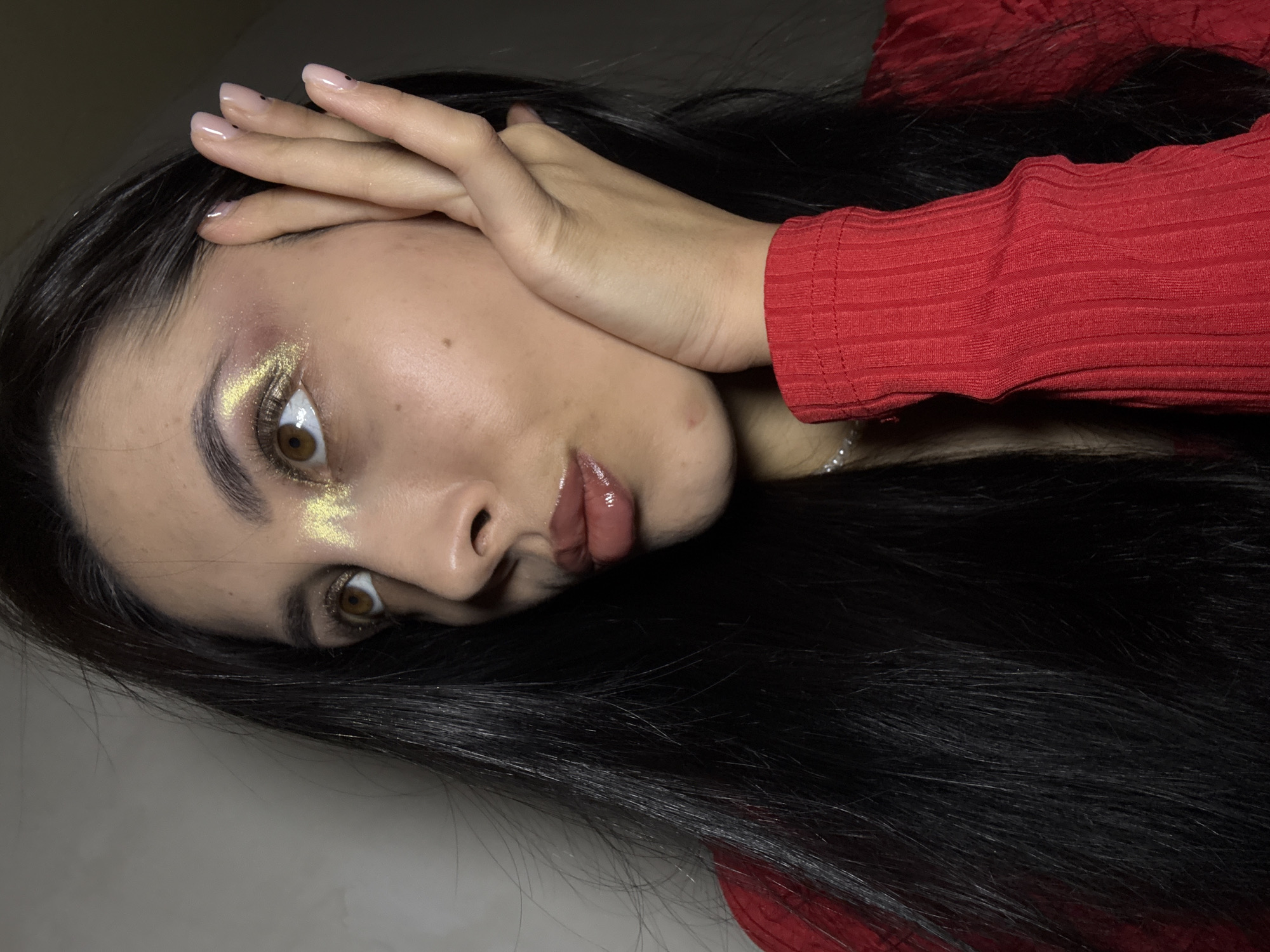 The image presents a close-up, tilted portrait of a young woman with long, dark hair. She looks directly at the viewer with intense brown eyes. Her right hand is raised, with manicured fingers resting gently on her forehead, partially covering her hair.
She is wearing a red, ribbed long-sleeved top or dress. Her nails are polished with a clear or light pink shade, and some fingers feature small, dark dot accents near the cuticles. Her makeup is a prominent detail, showcasing a generous application of shimmering golden glitter around her right eye, extending from the inner corner to the outer brow bone. Her lips are covered in a glossy, light brown or pink-toned product.
The background is a plain, light-colored surface, indicating an indoor setting. The lighting appears artificial, highlighting her features and the reflective glitter makeup. The woman's expression is neutral and posed, suggesting a self-portrait or fashion-oriented photograph. No specific activity is depicted beyond posing for the camera. There is no visible text in the image. This portrait is set in Votkinsk, Russia.
