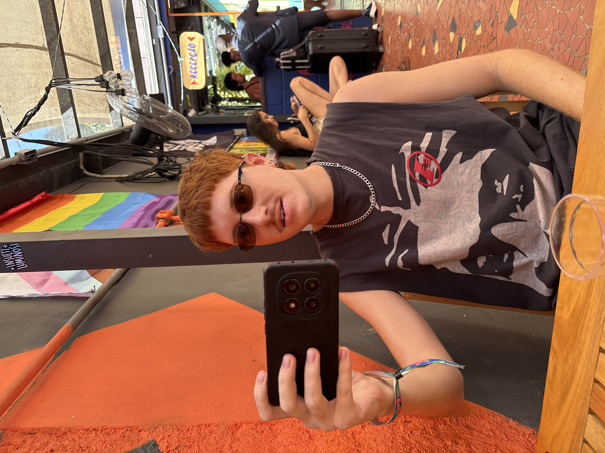 The image shows a selfie taken by a fair-skinned person with reddish-blonde hair, wearing sunglasses, a silver chain necklace, and a t-shirt with a large print on it. They are holding a black smartphone with four camera lenses. The foreground includes a partially visible drink in a glass.
In the background, several individuals are seated. One person has dark hair and is wearing a black top. Another person is lying down near a black suitcase.
A rainbow-colored flag is draped over a structure above the subject. Cables, lights, and a fan are also visible above.
A sign written in Portuguese ("RECEPCAO") is also visible. The image was taken in the city of São Paulo, Brazil.