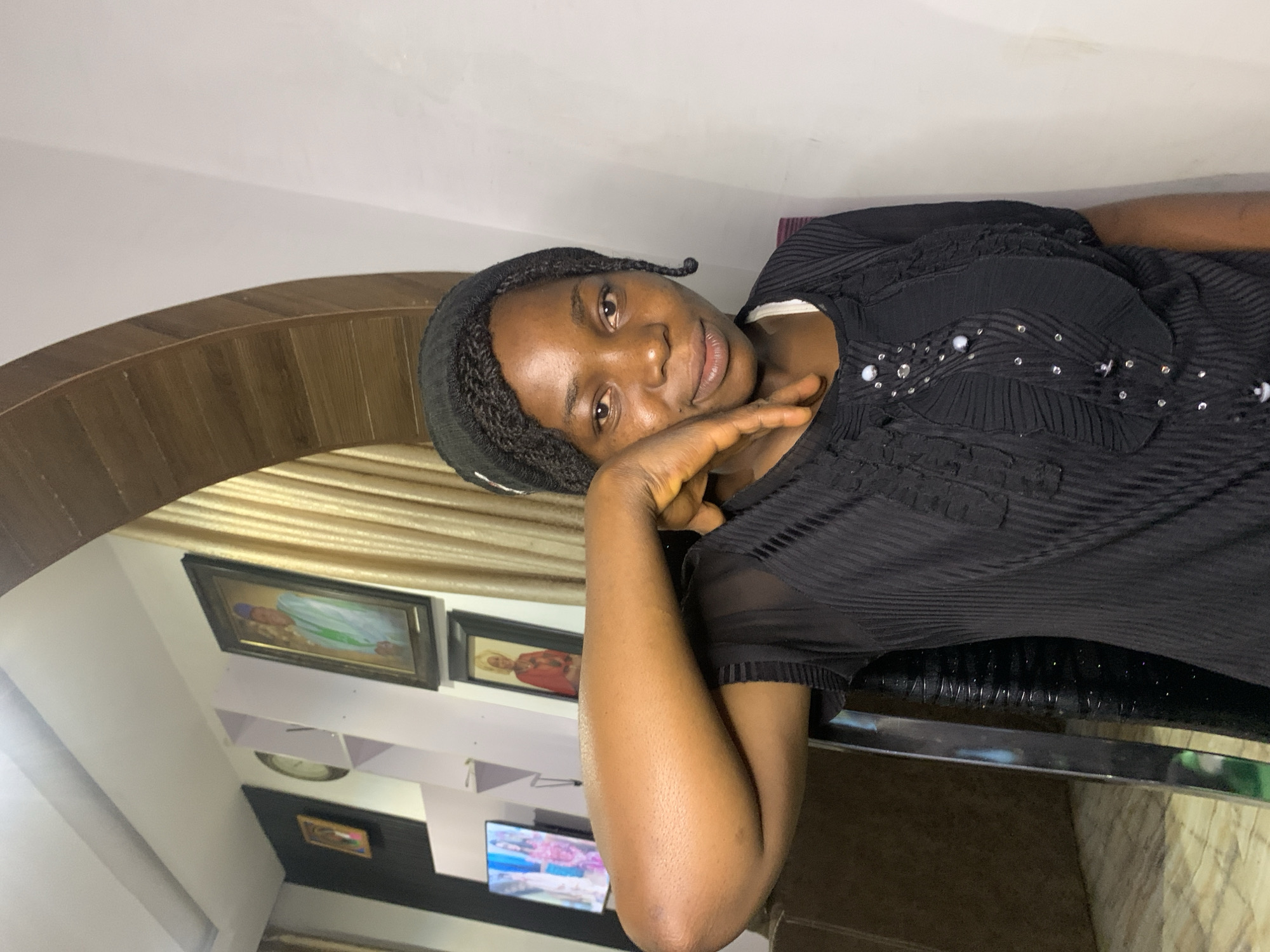 A young woman with dark skin and braided hair is seated in what appears to be a living room in Jos, Nigeria. She is wearing a black knit beanie and a black sheer top with decorative beading. Her right arm is bent, and her hand is resting on her cheek, giving her a thoughtful expression. The background features framed pictures on a wall, including portraits of people in traditional attire, and a glimpse of a couch. The room's decor suggests a modern but culturally rich environment.