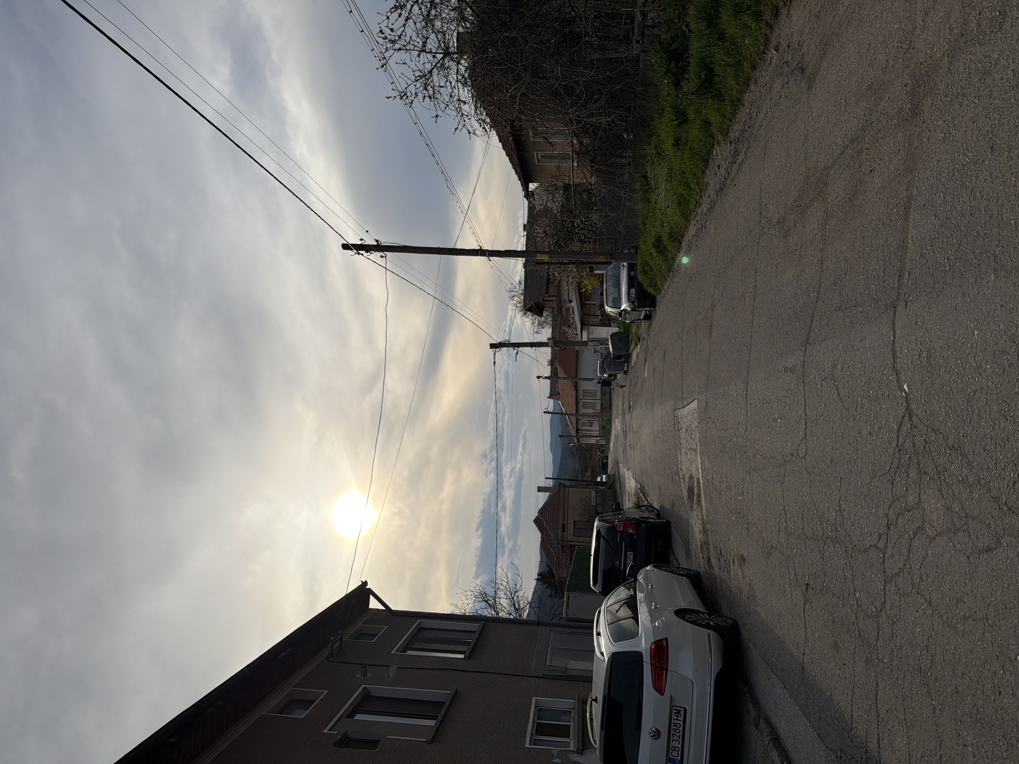 The image depicts a street scene in Popintsi, Bulgaria. The view is along a narrow asphalt road, showing cars parked on the right side. Buildings line the street, one of which is a two-story structure with visible windows. Overhead, power lines traverse the sky. The sky is overcast with visible sun. In the distance, the silhouette of hills can be discerned.