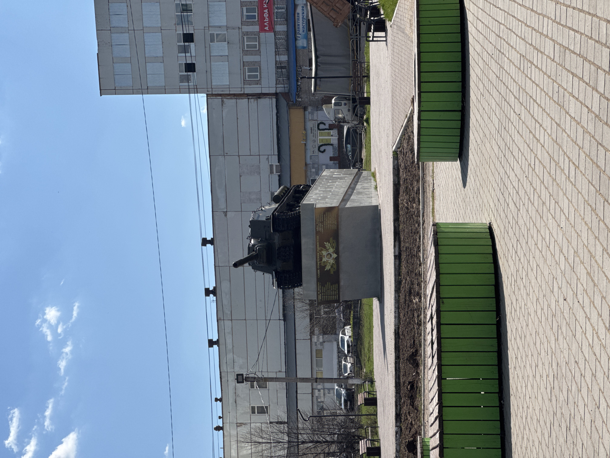 The image depicts a tank monument located in the city of Chaikovsky, Russia. The tank sits on a concrete pedestal with a plaque mounted on it, placed along a paved walkway lined with green benches. The backdrop includes a multi-story residential building with visible windows and a clear blue sky with scattered clouds. Overhead utility lines are also visible.