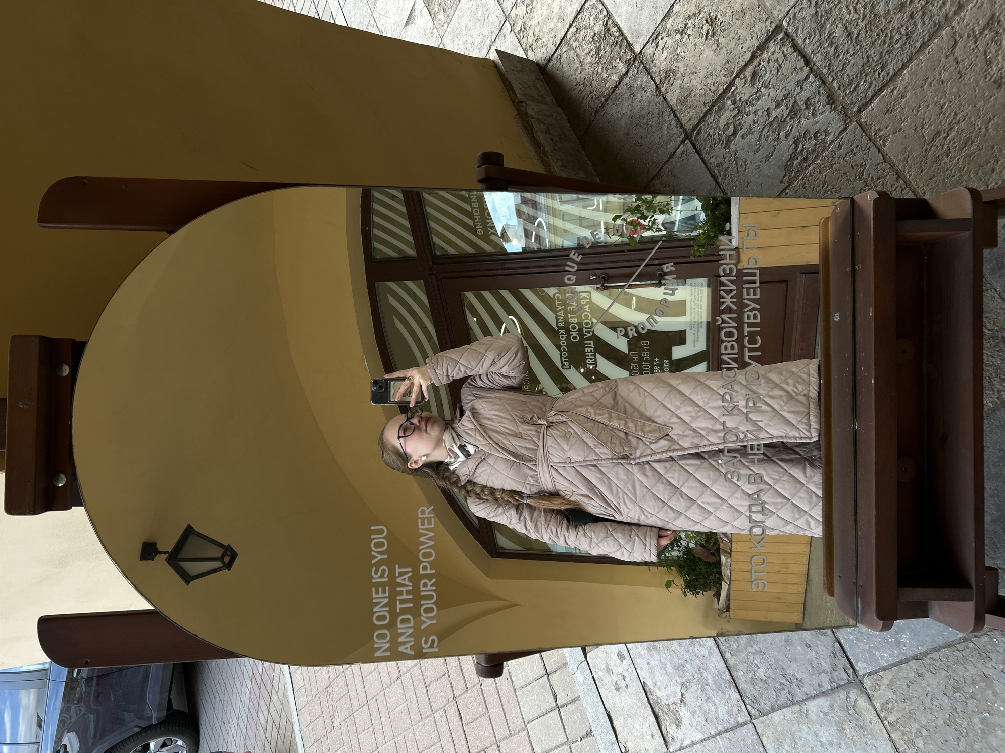 The image is a photograph of a girl taking a selfie in a mirror in Saint Petersburg, Russia. The mirror is oval-shaped and is mounted on a wooden stand. The mirror is situated on the sidewalk outside of a building with a brown door, and has lettering in both English and Russian. The English lettering is "NO ONE IS YOU AND THAT IS YOUR POWER". In the reflection, the girl is wearing glasses and a beige quilted coat and is holding a black smartphone up to take the photograph. The background includes grey stone pavement and a glimpse of a black car. The scene is situated in an urban environment.