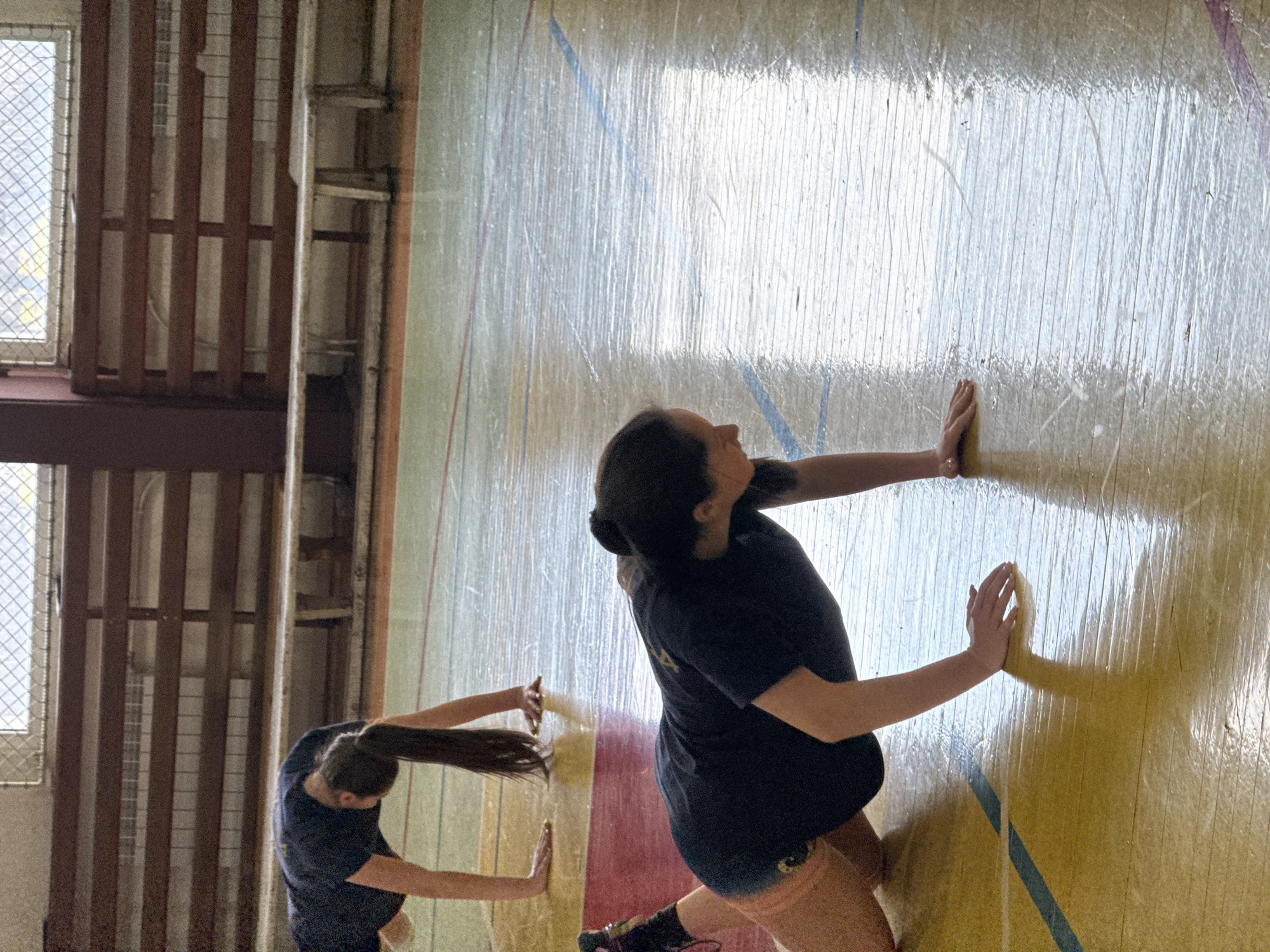 Two people are pressing their hands against a wall in what appears to be an indoor sports facility. Both are wearing dark T-shirts and shorts. One person has their hair pulled back in a ponytail. The other has their hair in a bun. The wall is glossy and marked with lines. There is a window with a metal grate and a section of wooden structure visible in the background. I cannot determine the location in Sofia, Bulgaria, from the image alone.